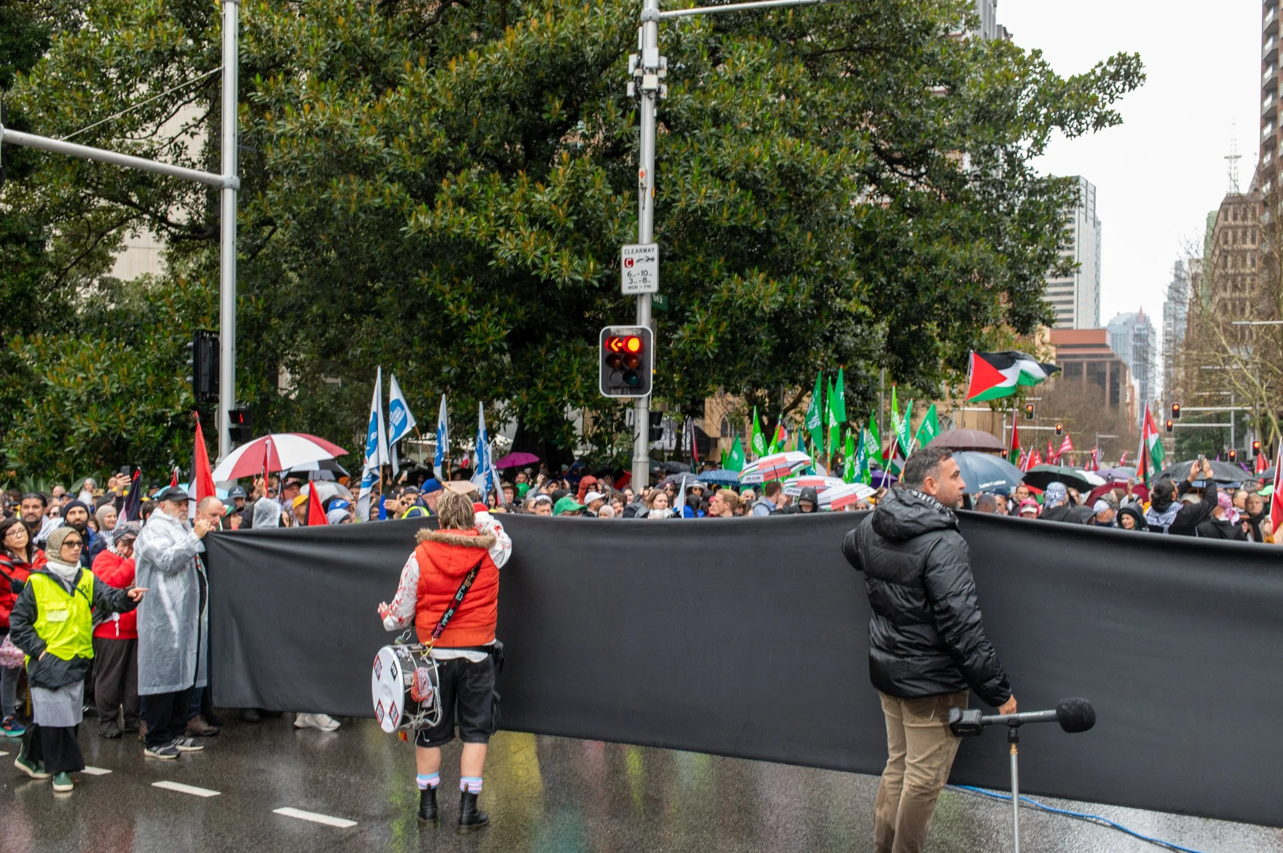 People participating in a protest or rally, holding signs and Palestinian flags, on a rainy day in an urban setting with tall buildings and trees in the background.