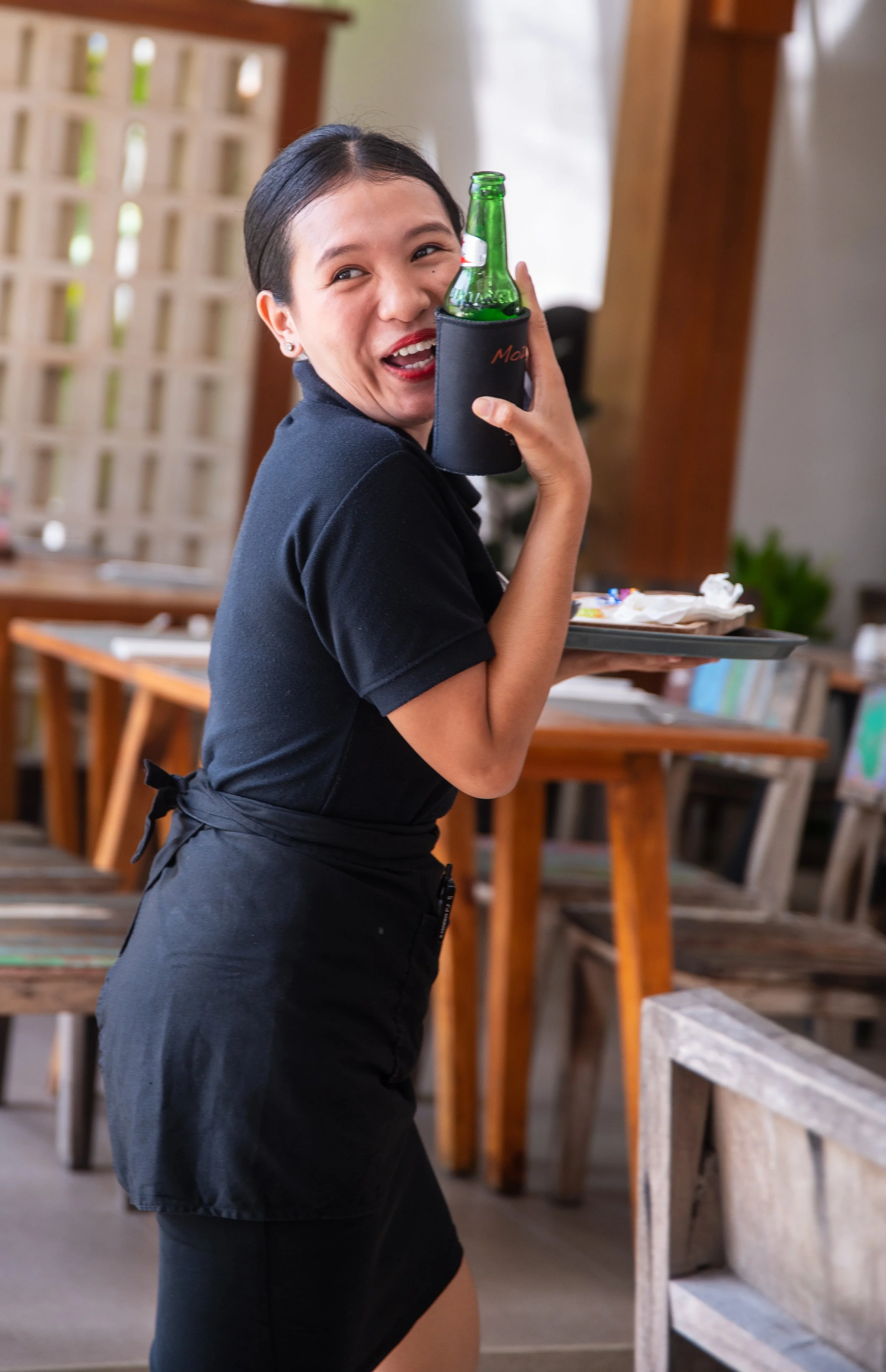 A woman in black clothing holding a green glass bottle in a black cozy holder, smiling, in a room with wooden furniture.