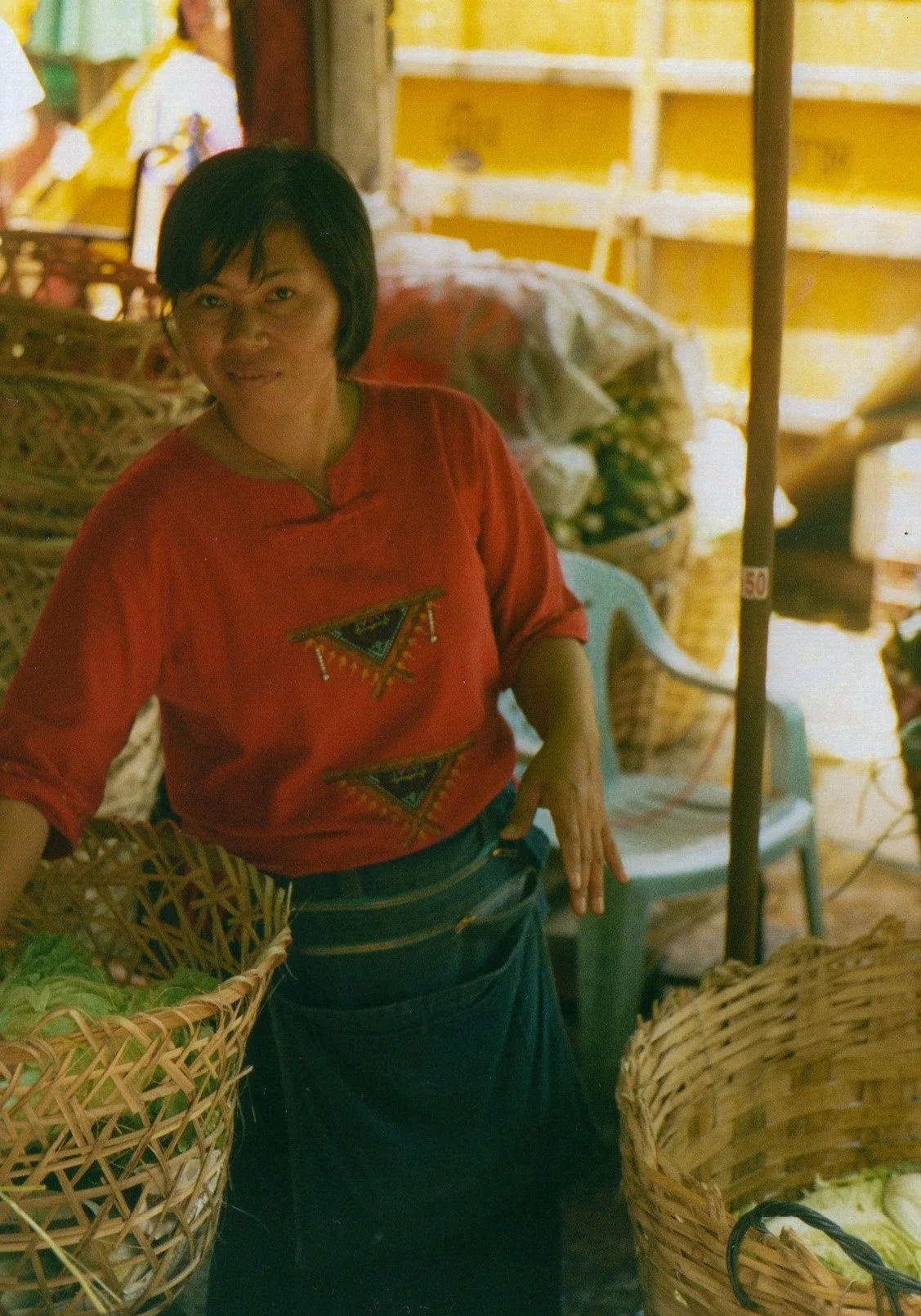 A woman wearing a red shirt and a dark skirt stands in a market stall with baskets of vegetables, smiling and posing for the photo.