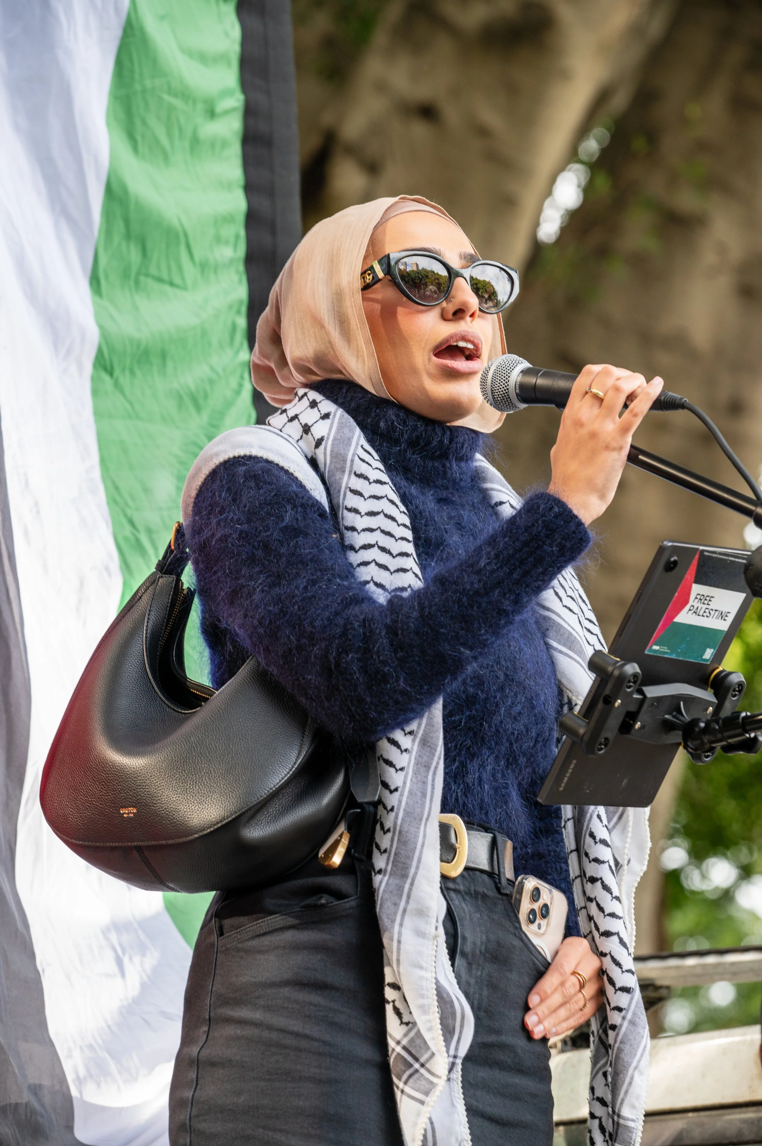 A woman wearing sunglasses and a beige headscarf speaking into a microphone during an outdoor event. She has a black handbag, is dressed in a navy blue fuzzy sweater, black pants, and has a phone in her pocket. She is standing in front of a tree and a banner with the colors green, white, and black.