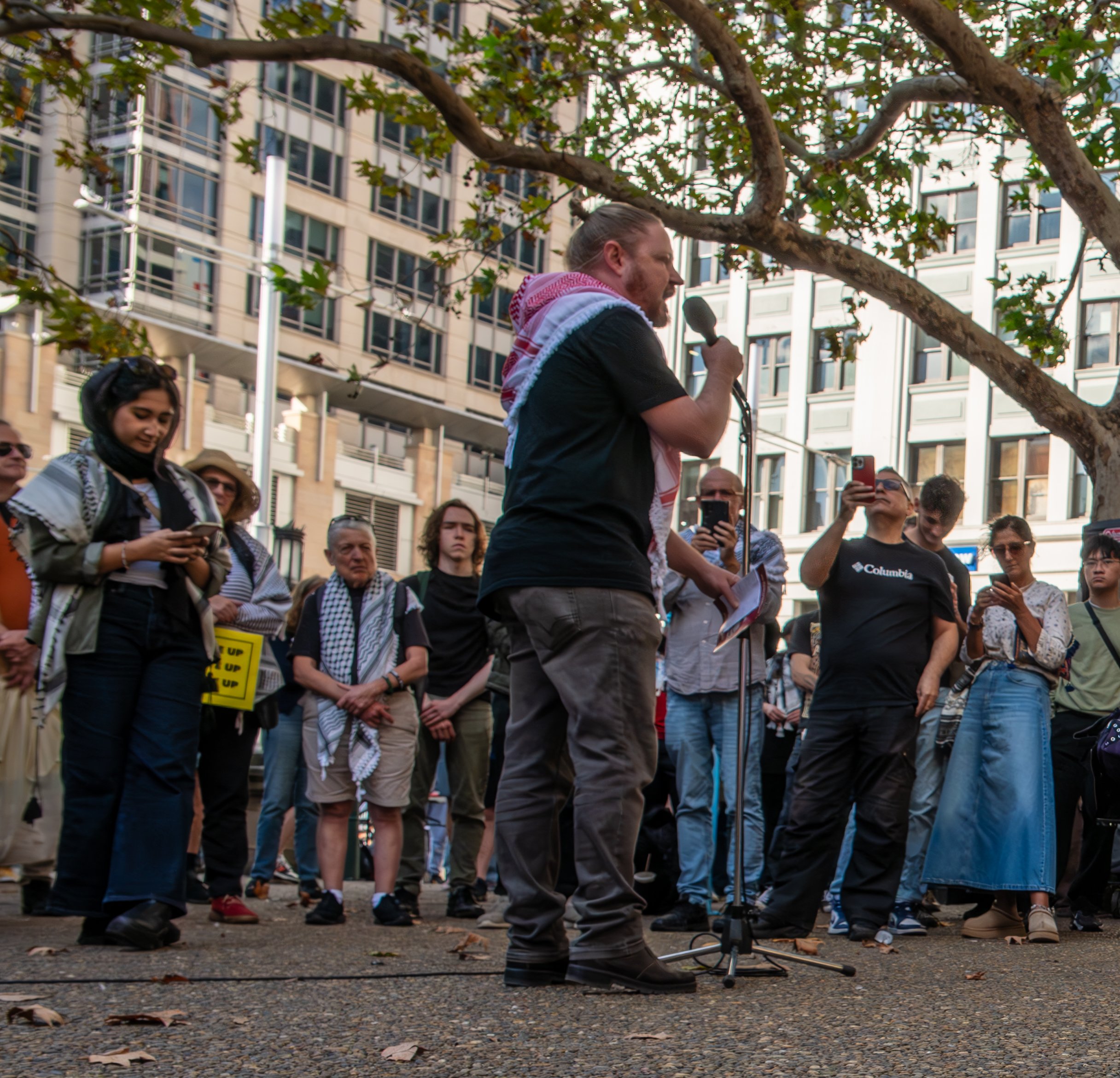 Man speaking into a microphone at a protest or rally, surrounded by a diverse group of people, some taking photos or listening, outdoors in an urban area with tall buildings and trees.