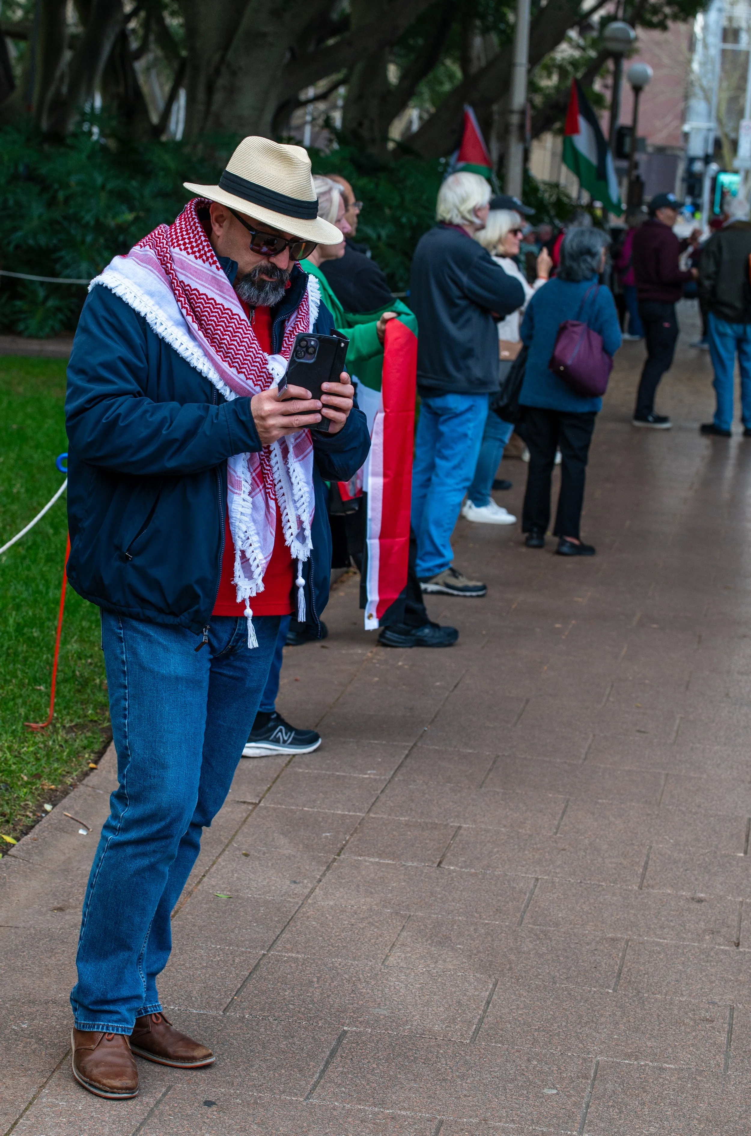 A group of people standing in line outdoors, with one man in the foreground wearing sunglasses, a straw hat, a scarf, and a navy jacket, looking at his phone. Others in line are holding flags and wearing casual clothing.
