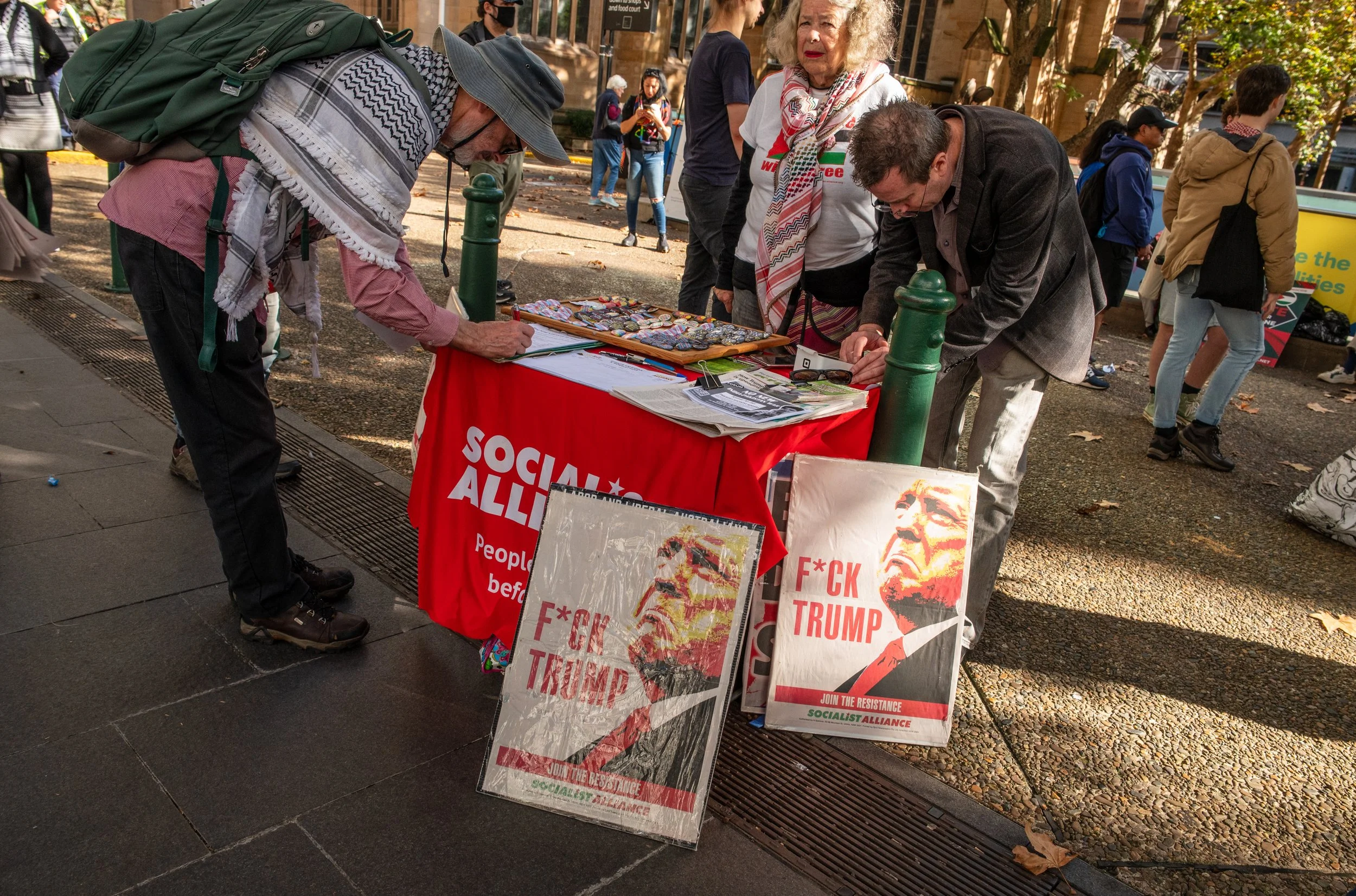People at a table with signs opposing Donald Trump, including one with his altered image and the text "F*CK TRUMP". The table has a Red cloth with white text reading "SOCIALISTS ALLIANCE". The scene is outdoors in a park or street, with other pedestrians in the background.