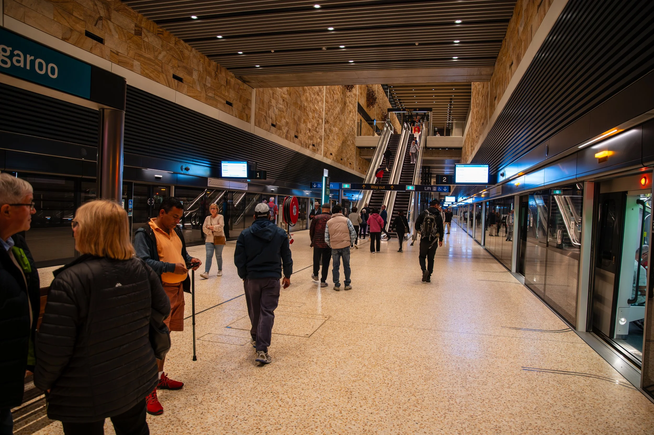 People walking inside a modern train station with escalators, digital signs, and glass doors, during daytime.
