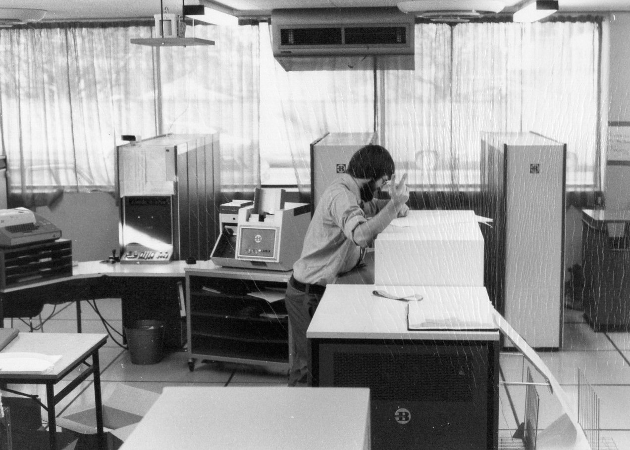 A man wearing a mask and glasses leaning over a desk with a large cardboard box, in an office filled with vintage office equipment, including printers, typewriters, and partitioned workspaces, with curtains covering large windows in the background.
