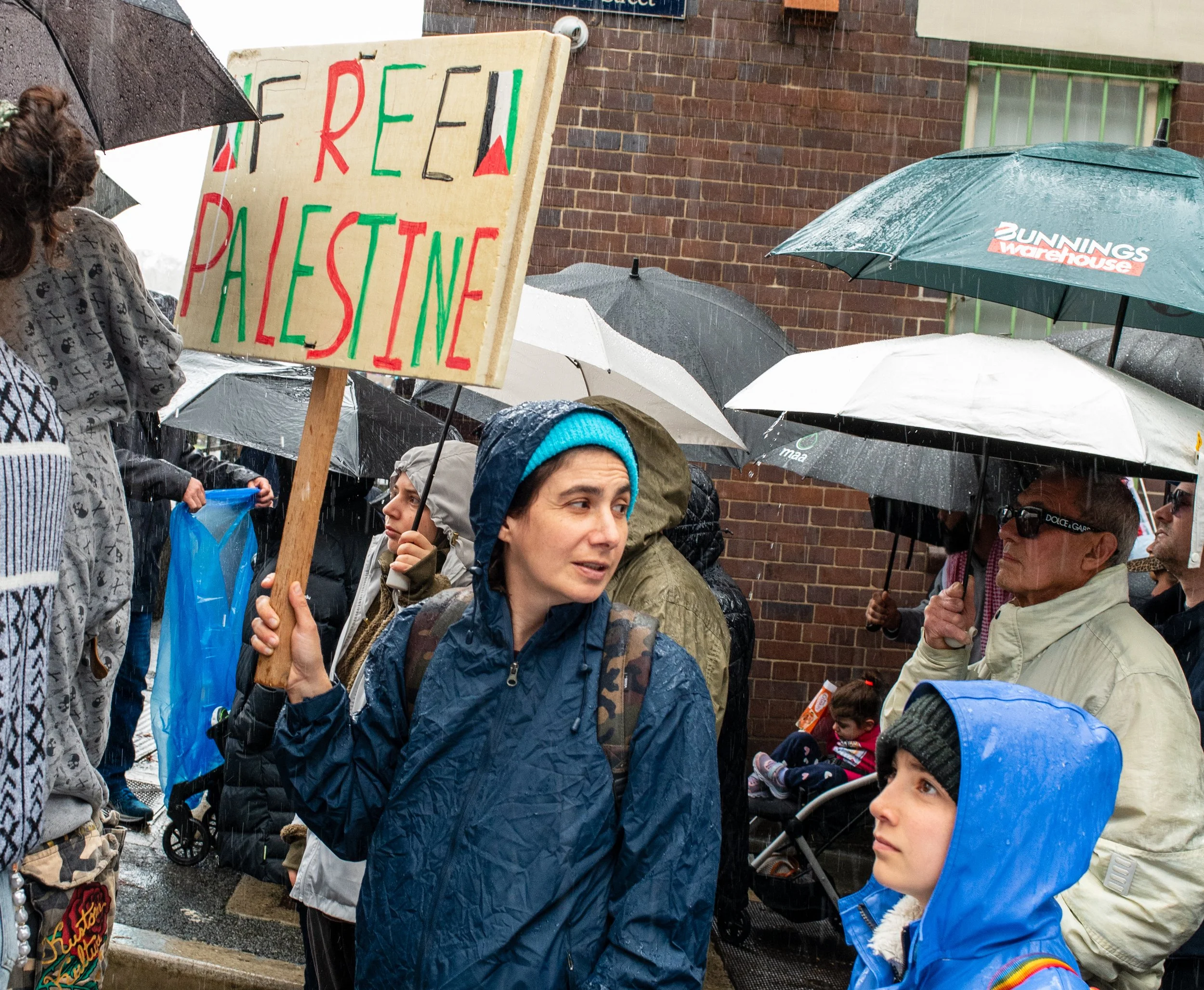 People holding umbrellas at a rain-soaked protest, including a woman holding a sign that reads 'FREE PALESTINE' in red and green letters.