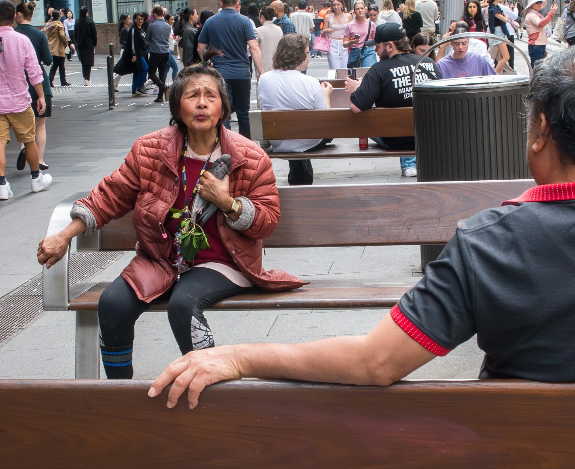An elderly woman sitting on a park bench with a surprised or expressive facial expression, gesturing with her left hand and holding a small plant in her lap, while a man sitting nearby has his arm stretched along the backrest. The background is a busy city street with numerous pedestrians walking and sitting.
