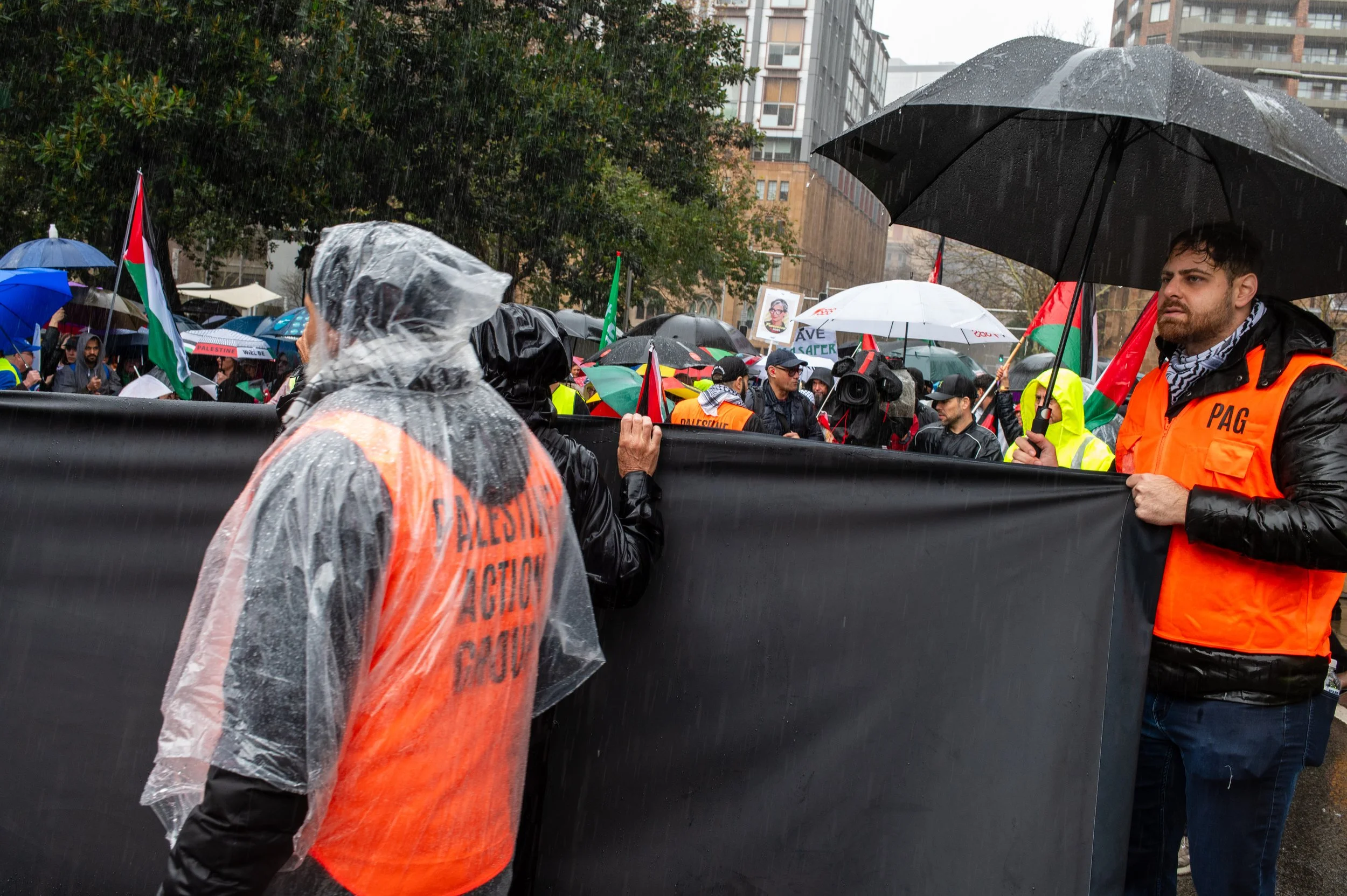 A crowd of protesters holding flags and banners in rainy weather, some wearing rain ponchos and orange vests, standing behind a large black barrier in an urban setting.