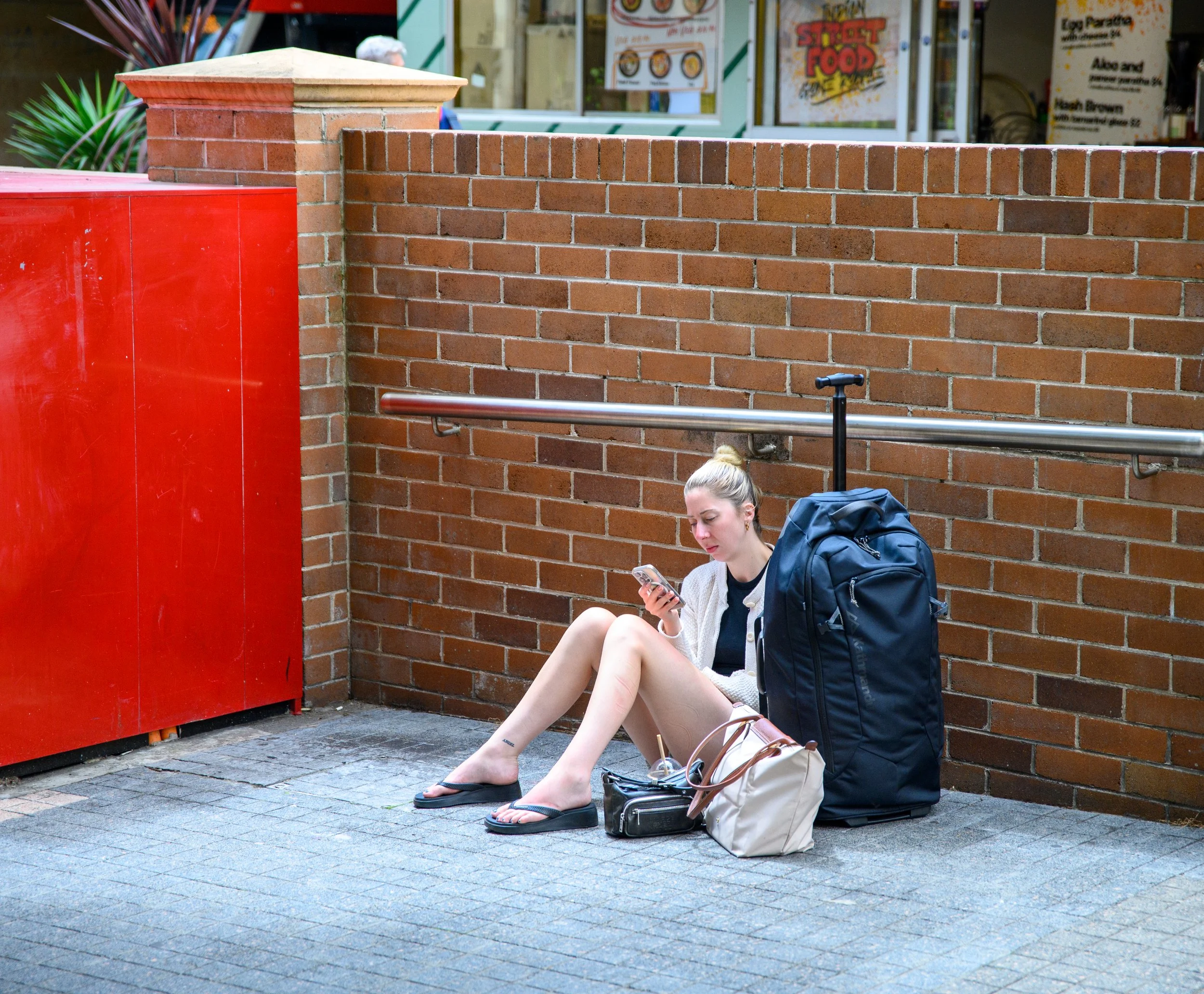 A woman sitting on the sidewalk against a brick wall, looking at her phone, with several luggage and bags beside her.
