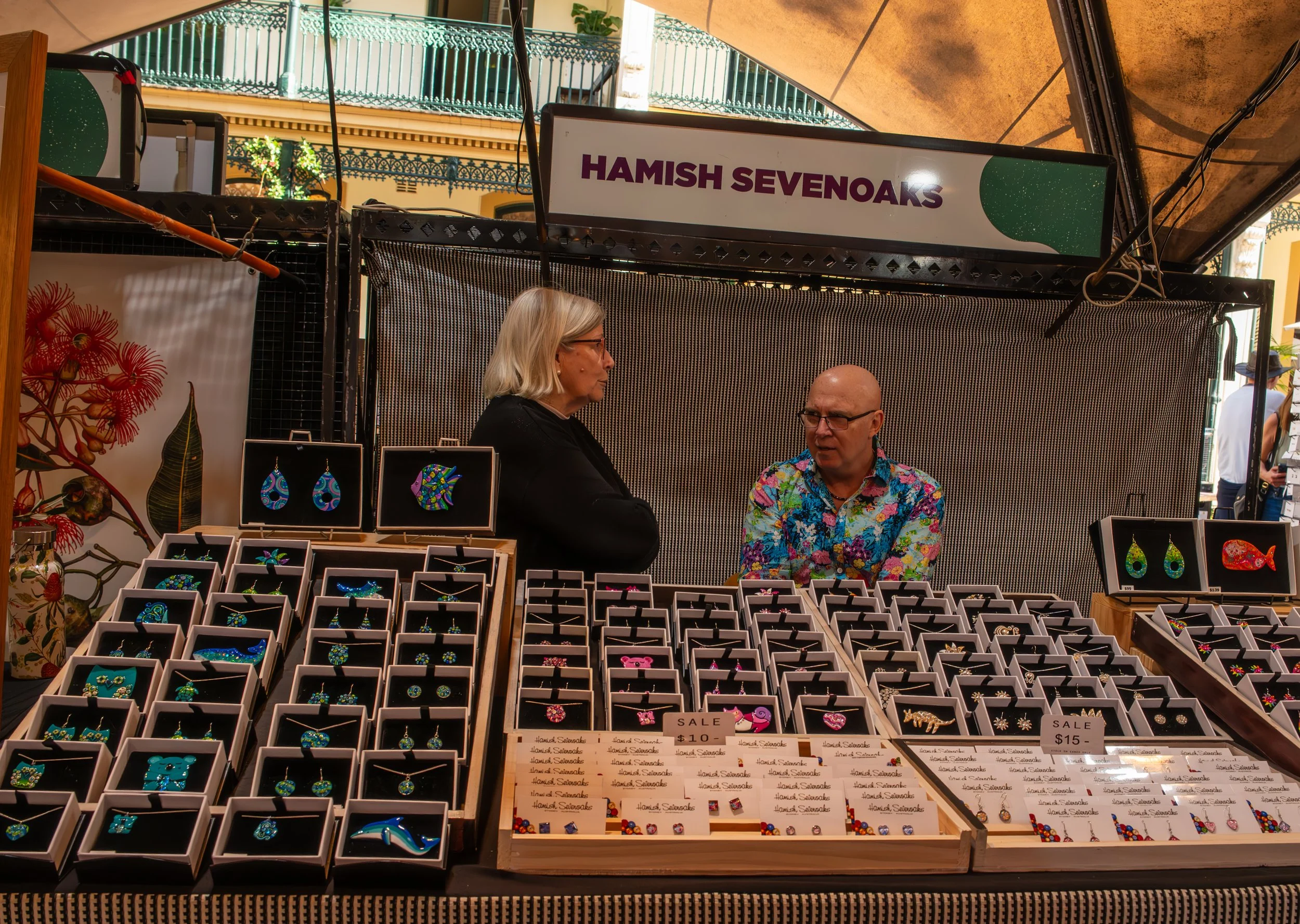Jewelry vendor at market stand with colorful earrings and necklaces, with two women talking, one with glasses and gray hair, and the other bald, wearing a floral shirt, at the Hamish Sevenoaks stall.