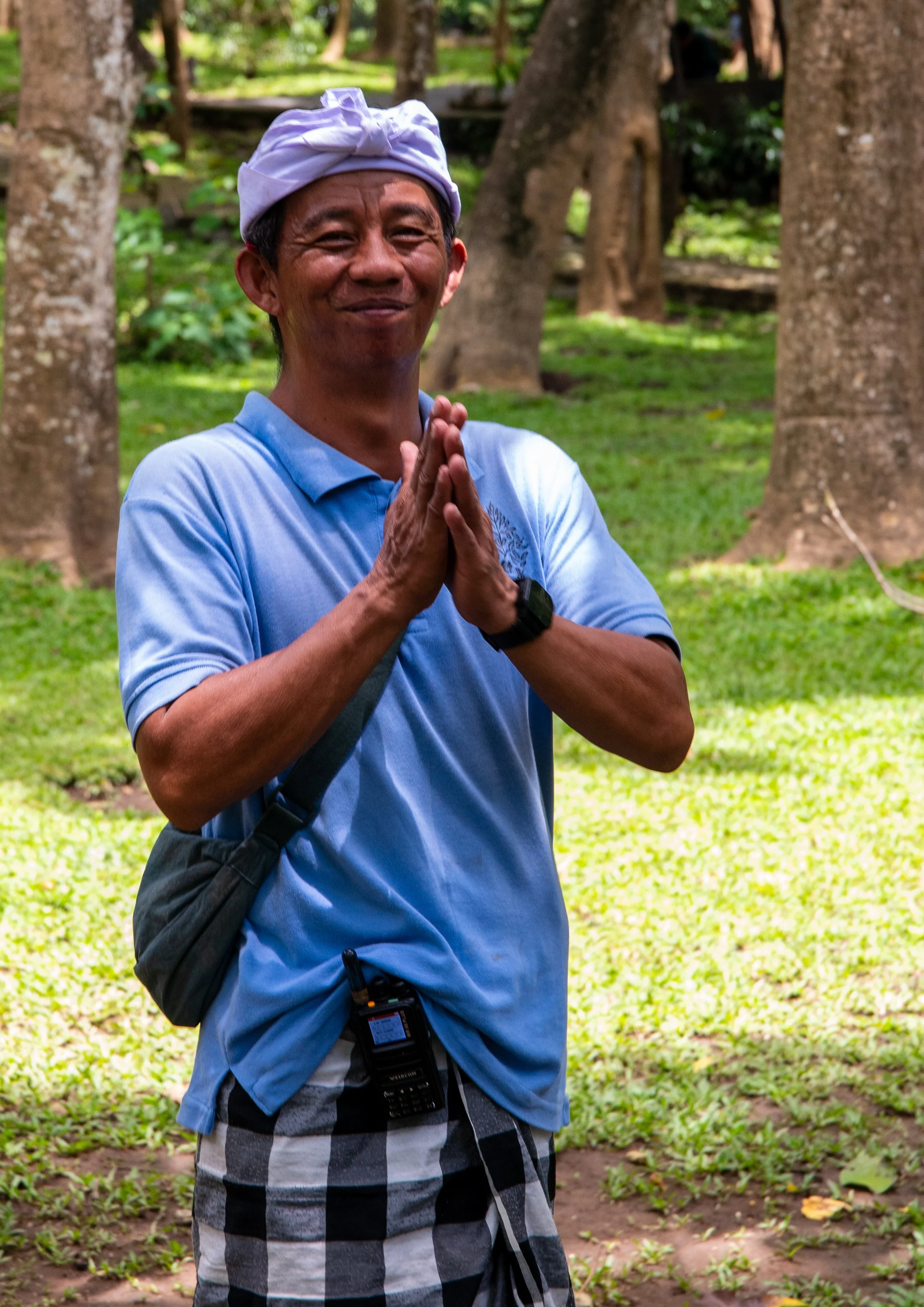 A smiling man wearing a white traditional headband, blue polo shirt, and plaid shorts stands outdoors in a lush green park with trees and grass, with his hands pressed together in a gesture of greeting or gratitude.