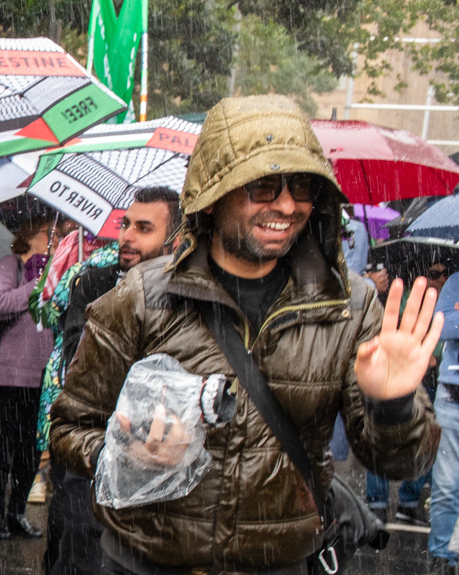 Man in military-style hat and sunglasses smiling and waving in the rain, holding a plastic water bottle, with a crowd in the background holding umbrellas.