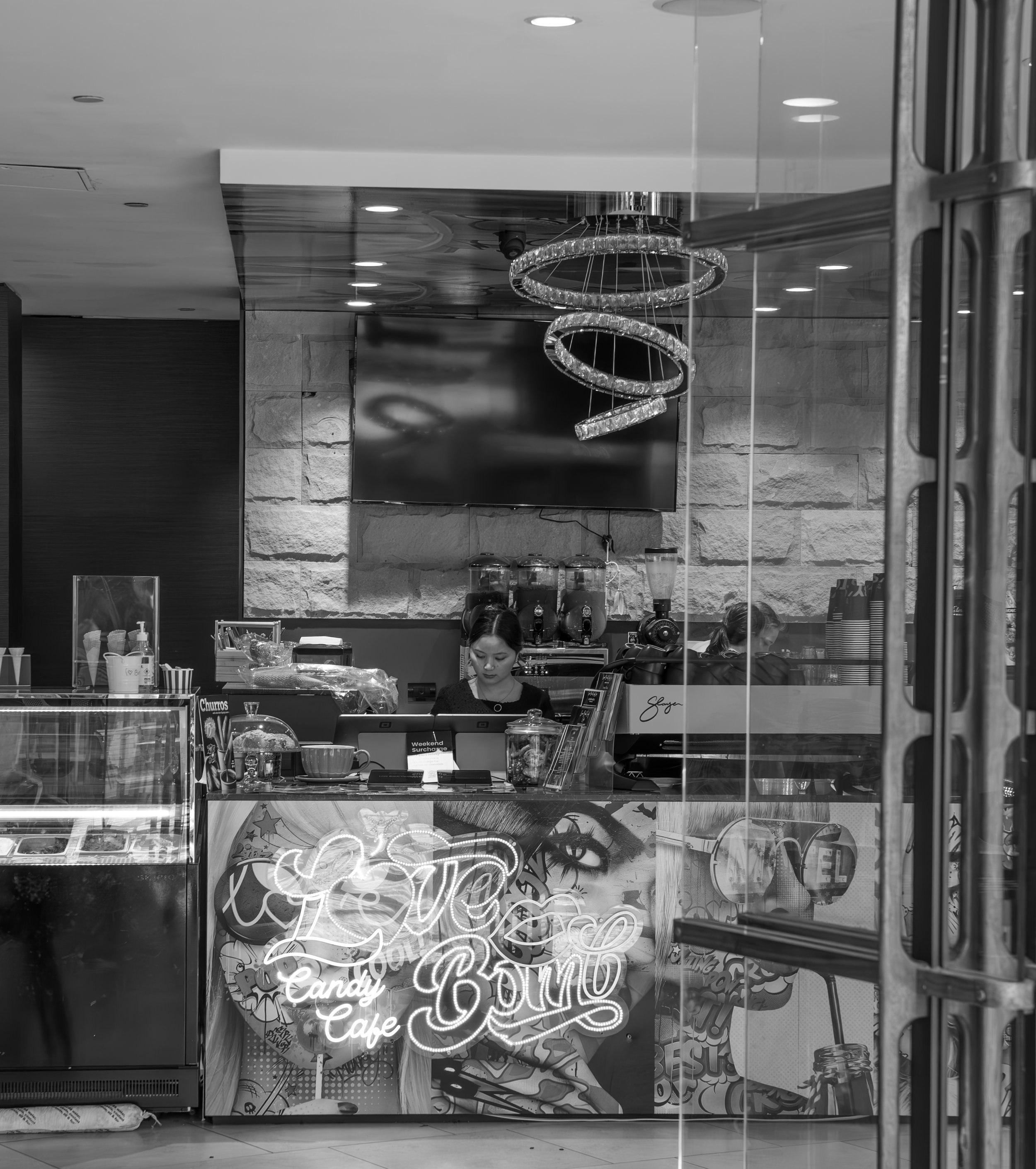 Black and white photo of a girl working at the counter of a candy cafe called Love Bomb, with a neon sign, graffiti art, and modern decor inside.