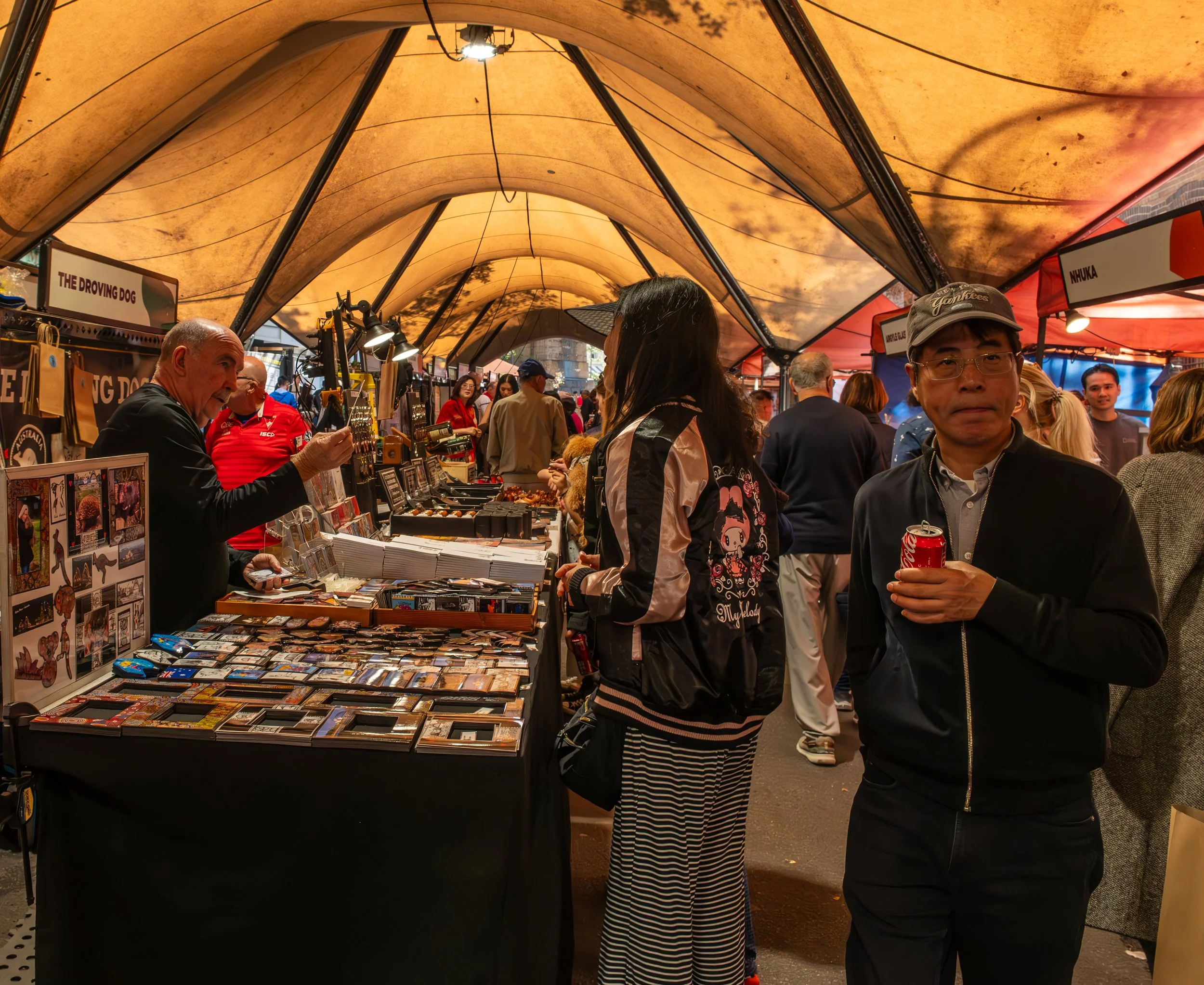People shopping at an outdoor market stall under a canopy, browsing various items and holding drinks.