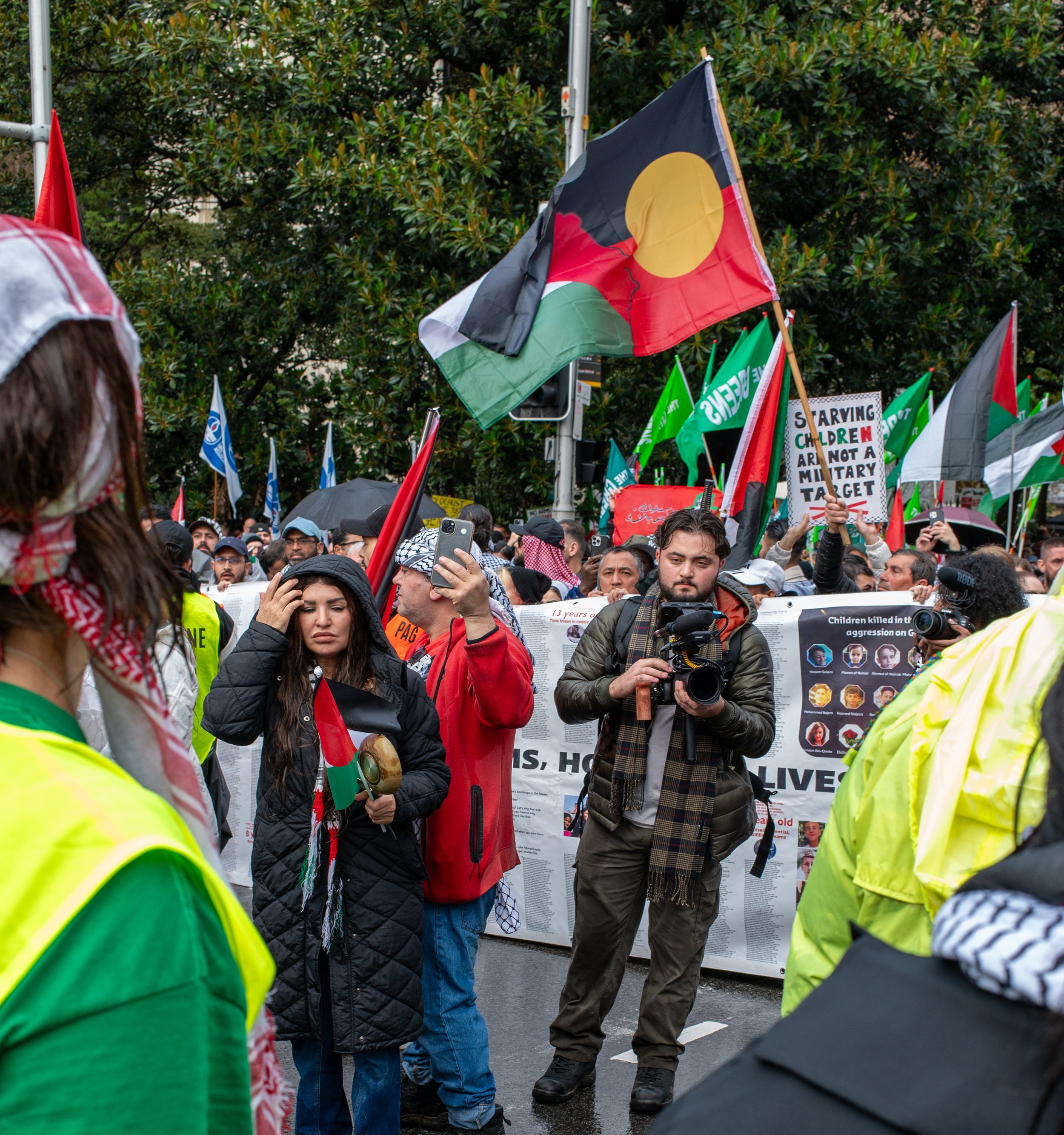 A group of people protesting show flags, signs, and a camera operator, with a green leafy background. Some protesters hold flags, including one with black, red, green, and yellow colors, and signs advocating for children and against military targets.