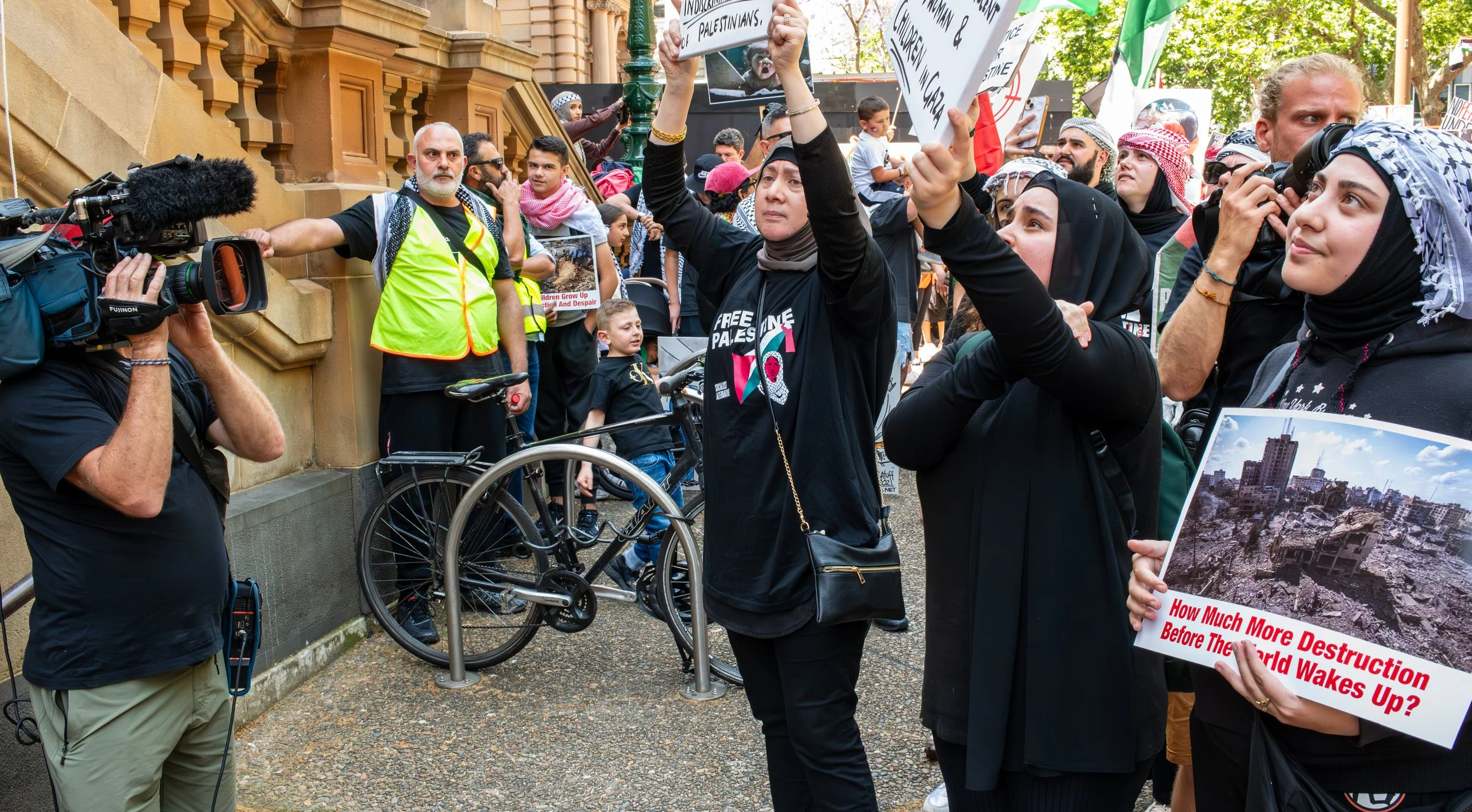 Protesters holding signs and banners at a rally, including one with an image of destruction and a caption questioning future devastation, with a crowd of people and a cameraman recording the scene.