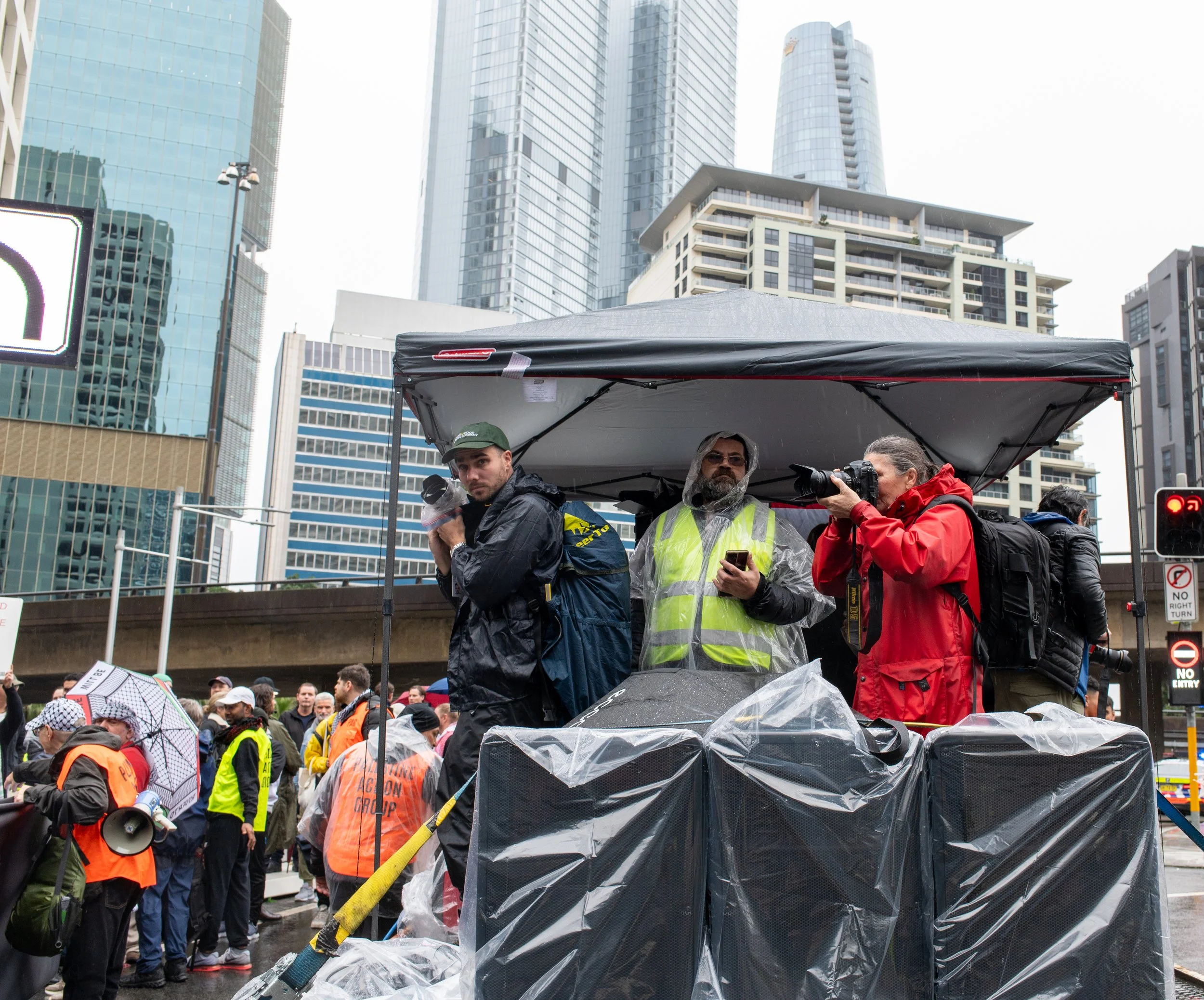 People gathered at a protest or rally in an urban setting with tall buildings, some wearing rain gear and holding umbrellas. Three individuals are under a temporary shelter: one holding a camera, one looking at a device, and another with a camera. Many participants in the background wear orange vests, and there is a crowd behind them.