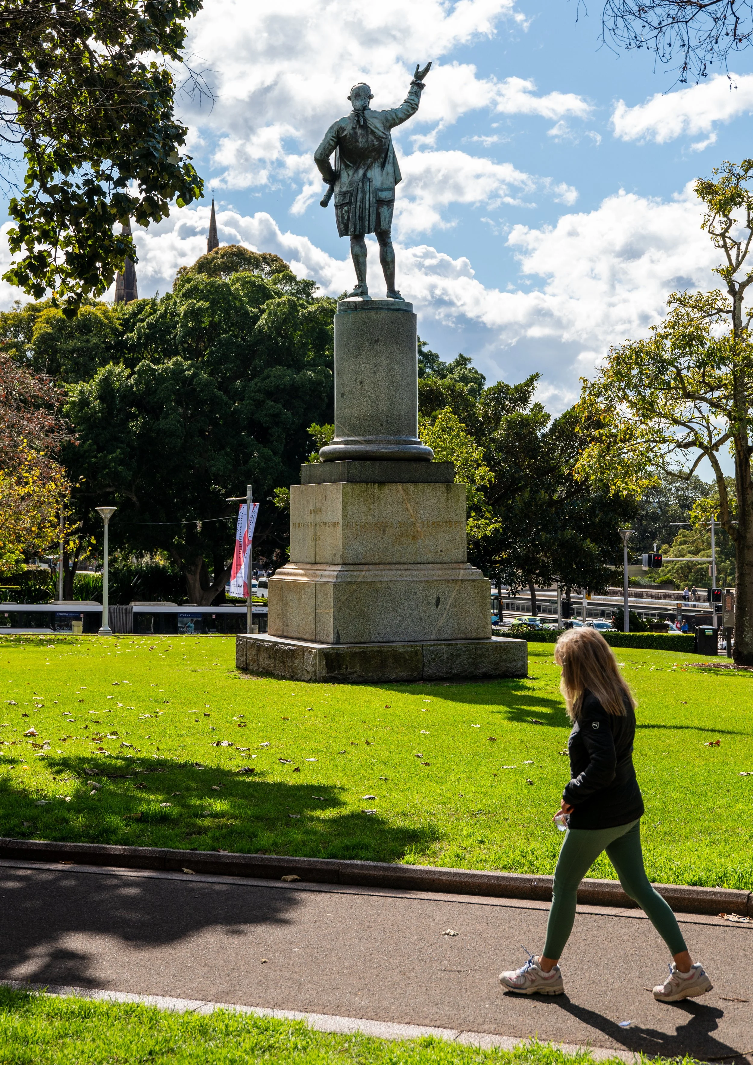 A woman walking past a bronze statue of a man on a pedestal in a park with green grass, trees, and cloudy blue sky.