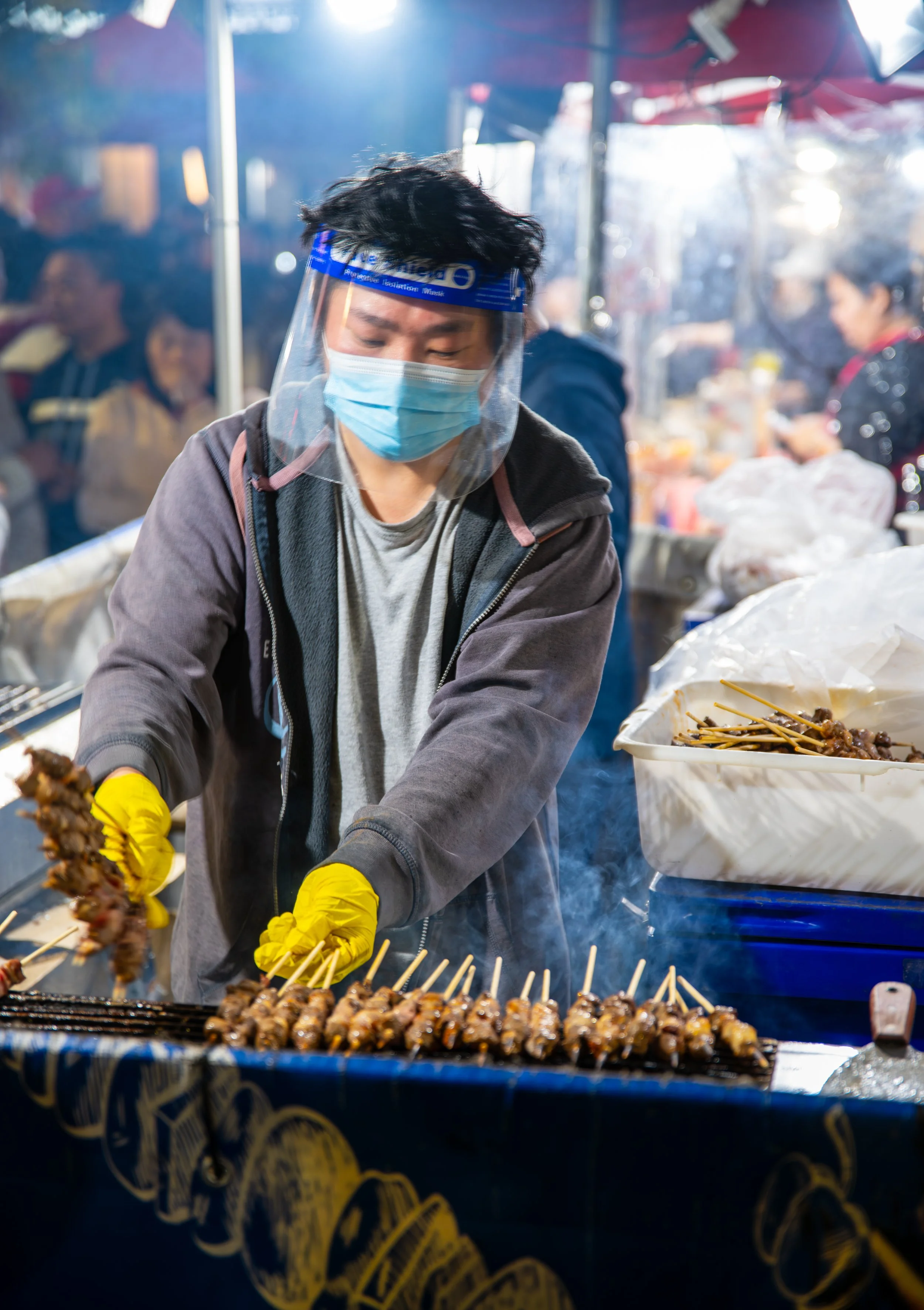 A person wearing a face mask and face shield cooking skewered meat at a street food stall in an outdoor market at night.
