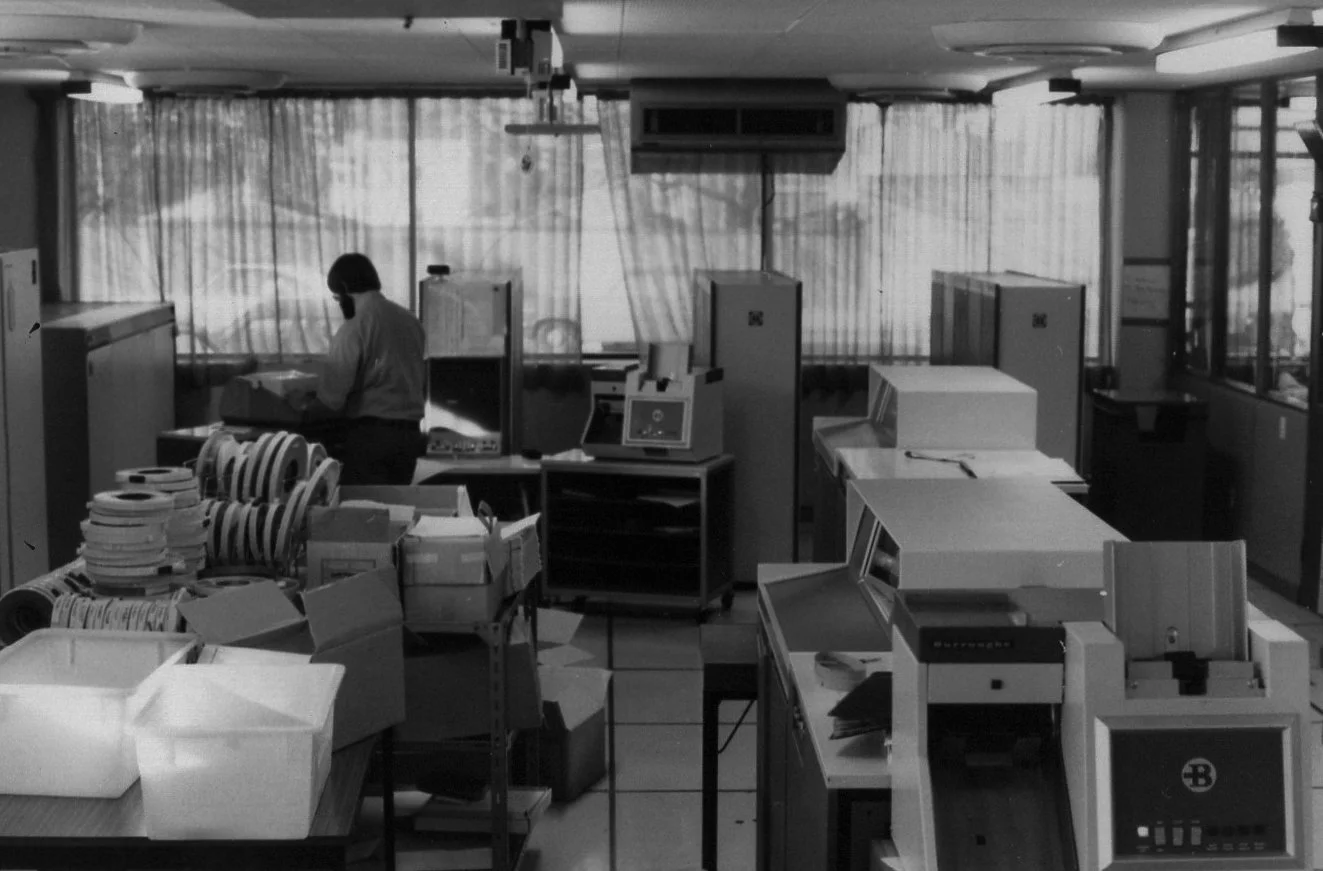 An office with desks and equipment, a person working near the window, various boxes, and office machines in black and white.