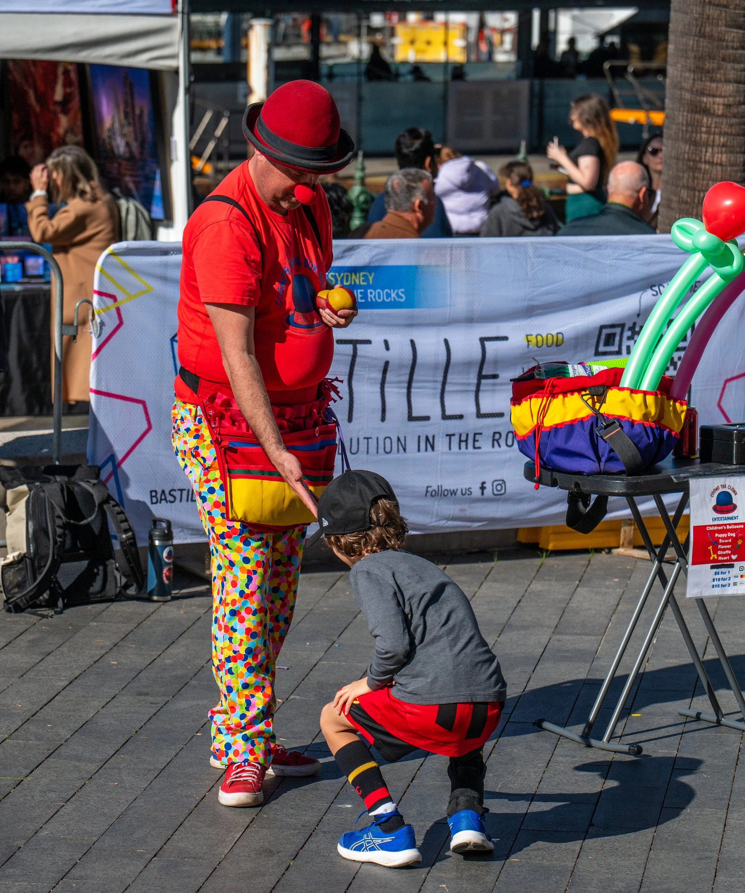 A clown in red, yellow, and polka dot clothing standing on a boardwalk, holding balloons, talking to a young boy who is crouching down. There are booths and people in the background.