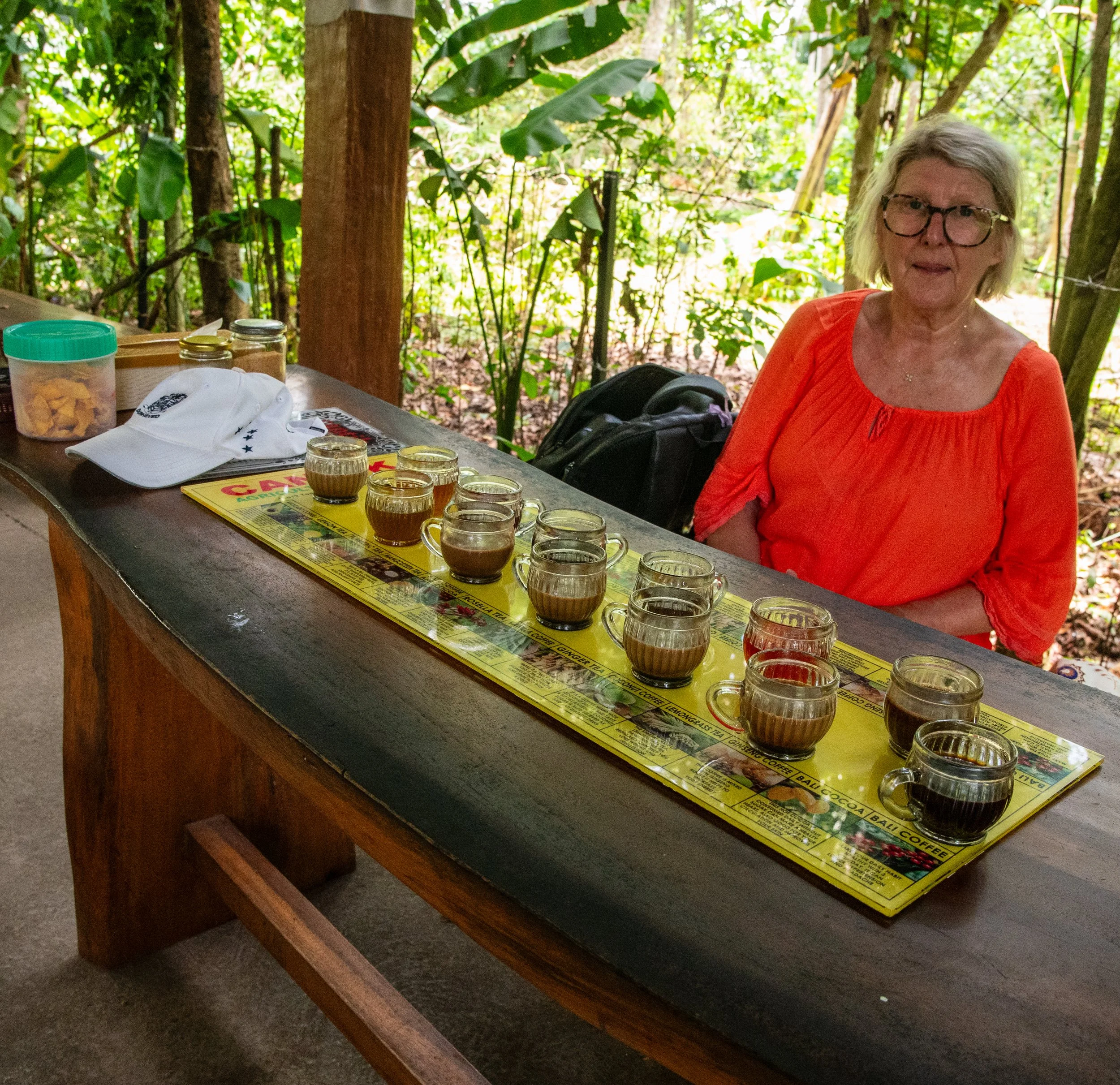 A woman with blond hair, glasses, wearing an orange top, seated outdoors at a table surrounded by green trees.