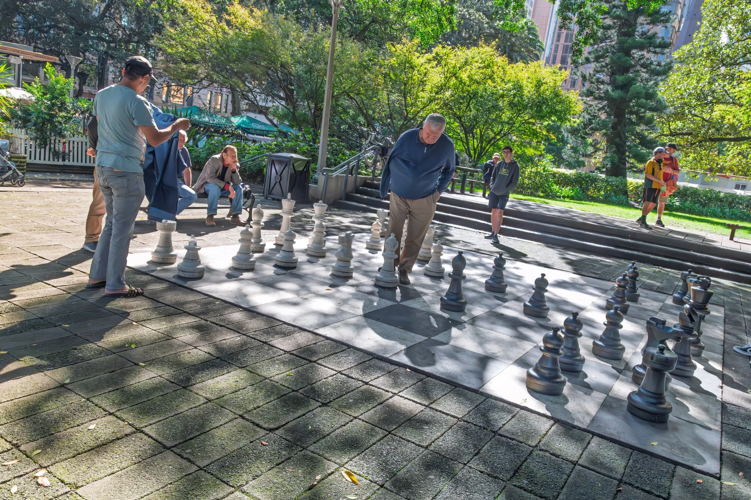 People playing chess on an outdoor chessboard in a park with trees and buildings in the background