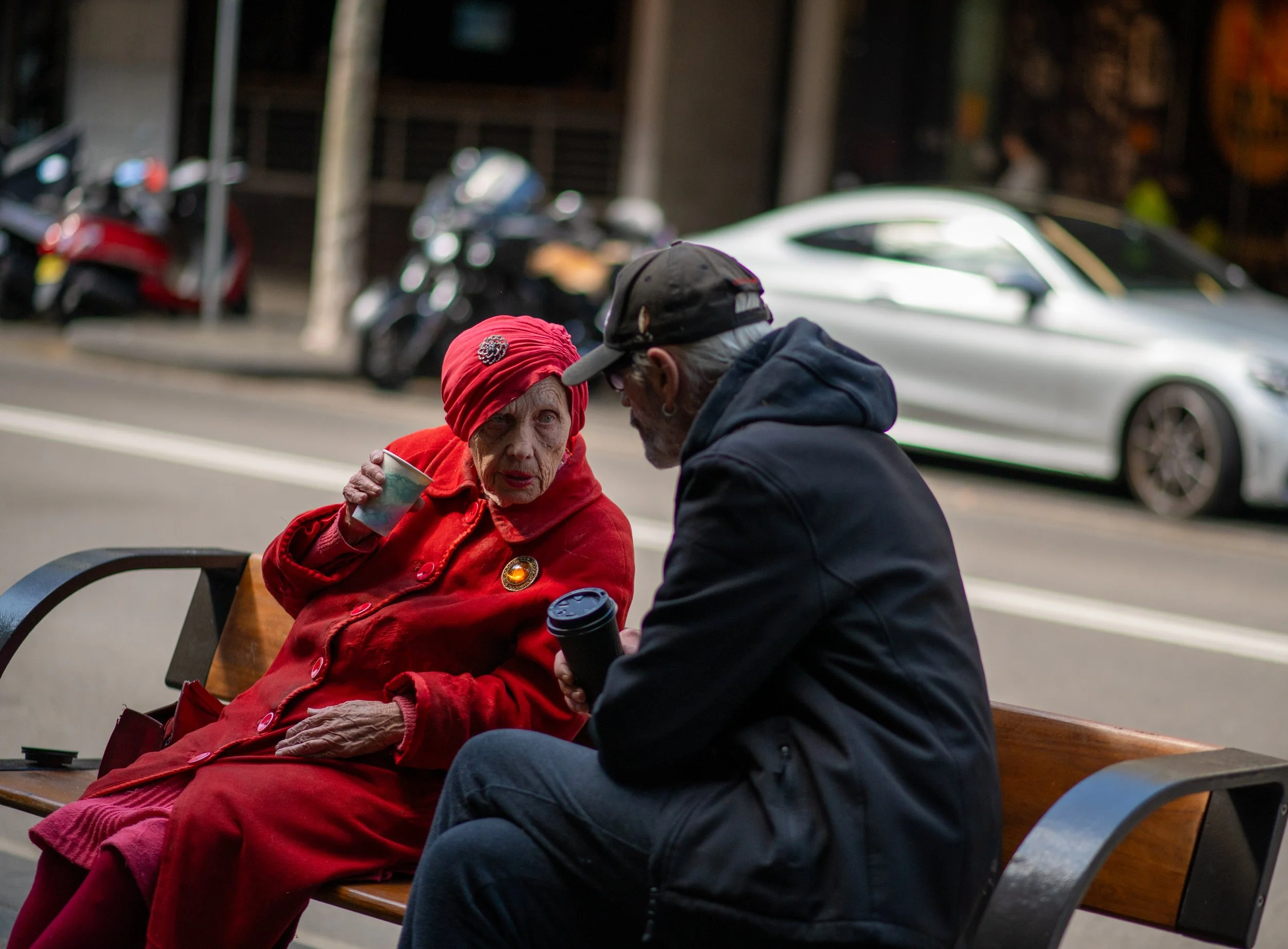 An elderly woman in a red coat and headscarf sitting on a bench talking to a man in a black hoodie and cap holding a coffee cup on a city sidewalk with cars and motorcycles in the background.