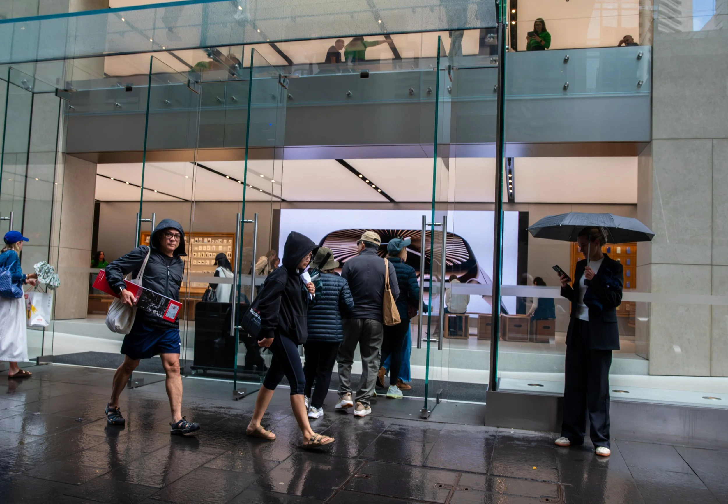 People entering an Apple store on a rainy day, with some carrying umbrellas and wearing rain jackets, and others walking on wet pavement.