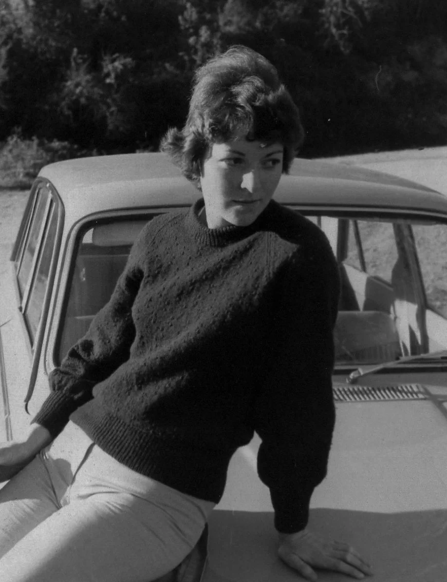 A black and white photo of a woman sitting on a car hood outdoors, dressed in a dark sweater and light pants, with trees in the background.