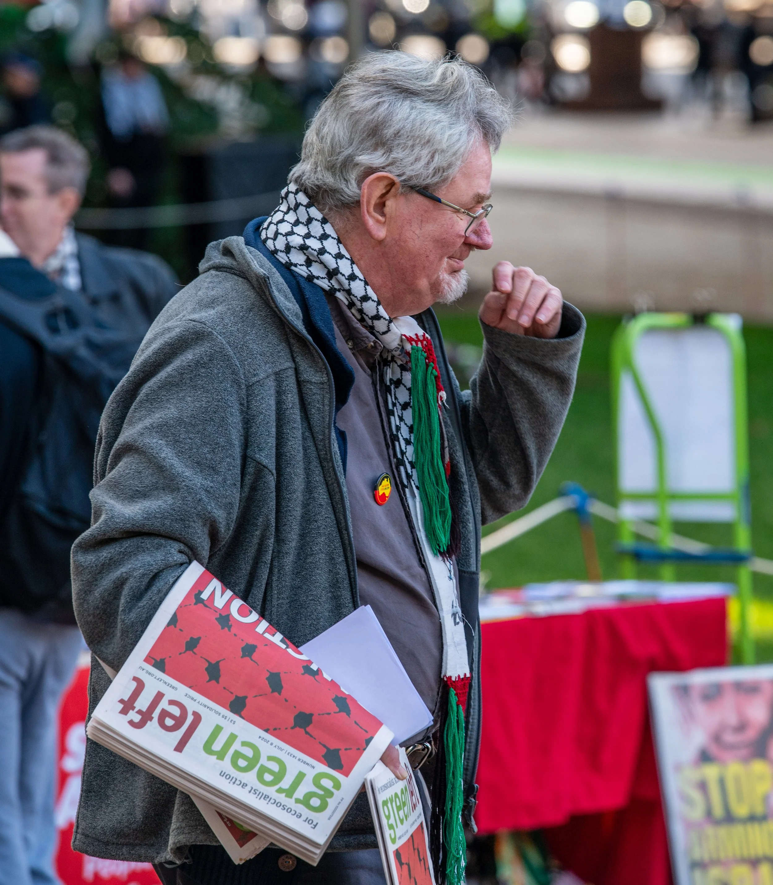 A man in a gray hoodie, wearing a black and white scarf, holding a stack of Green left newspapers at a protest or rally. He appears to be crying or emotional.