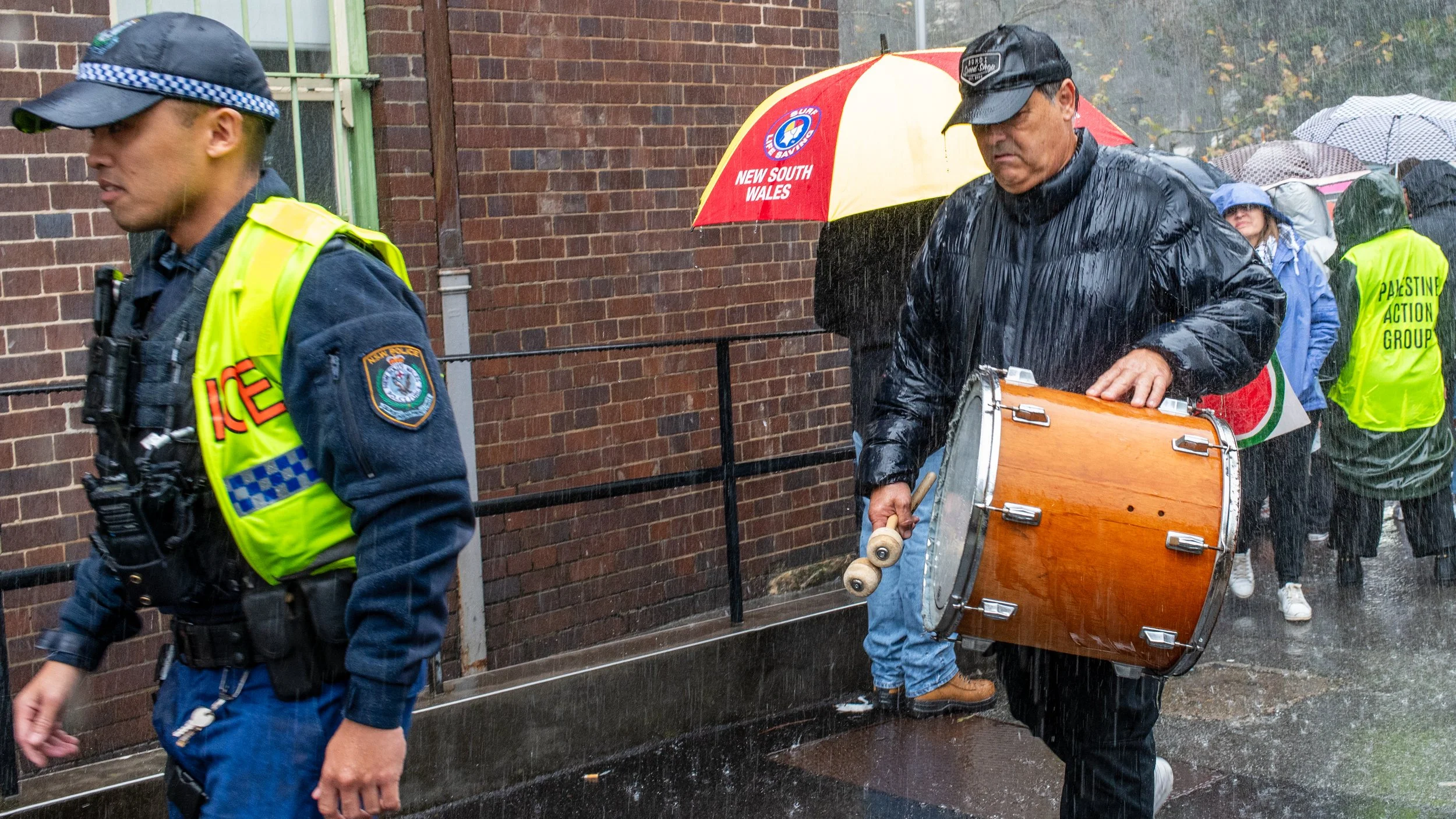 Police officer and man with drum walking in the rain, other people holding umbrellas in the background.