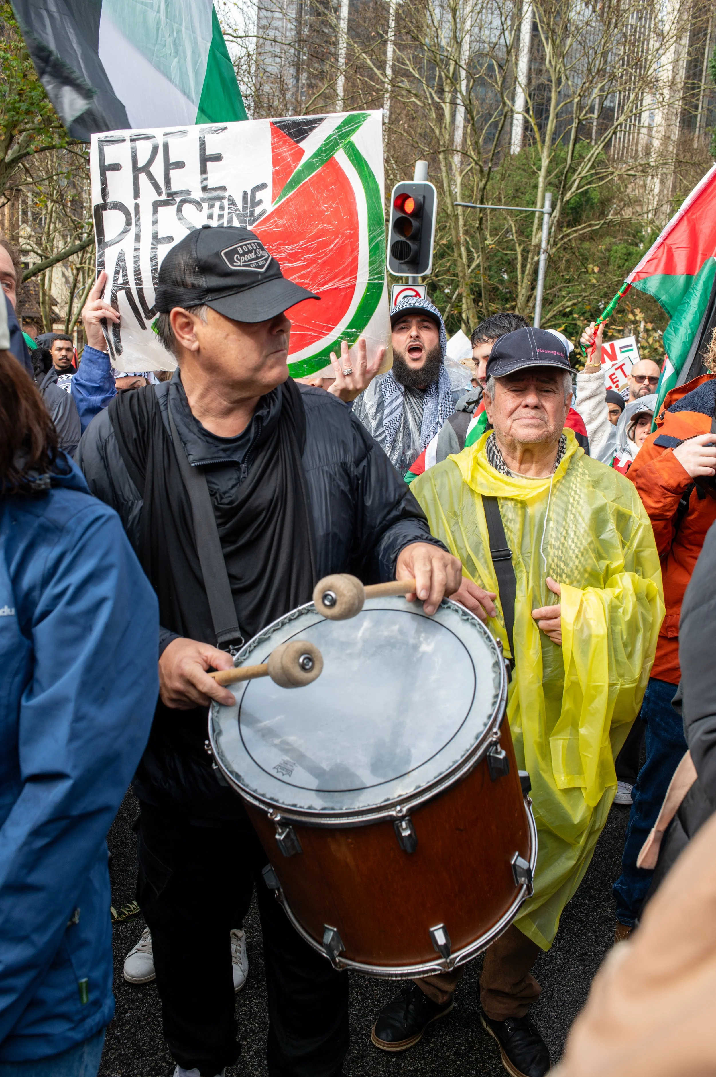 A group of people participating in a protest or demonstration outdoors, with some holding flags and signs. One person is playing a drum while others are gathered around, wearing raincoats and hats.