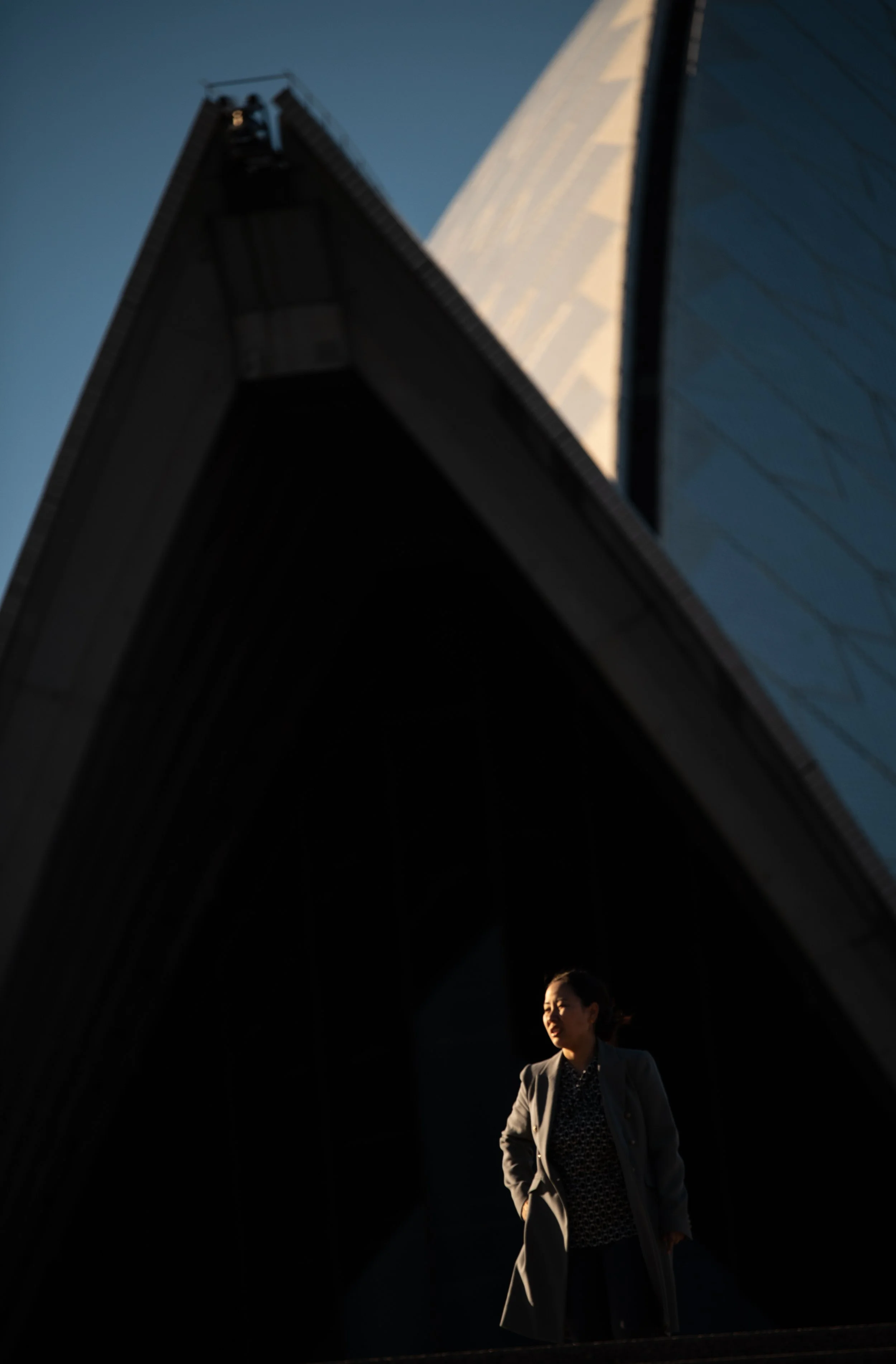 A woman in a gray coat standing in shadows beneath a modern architectural structure with large, pointed edges and reflective surfaces, with a clear blue sky in the background.