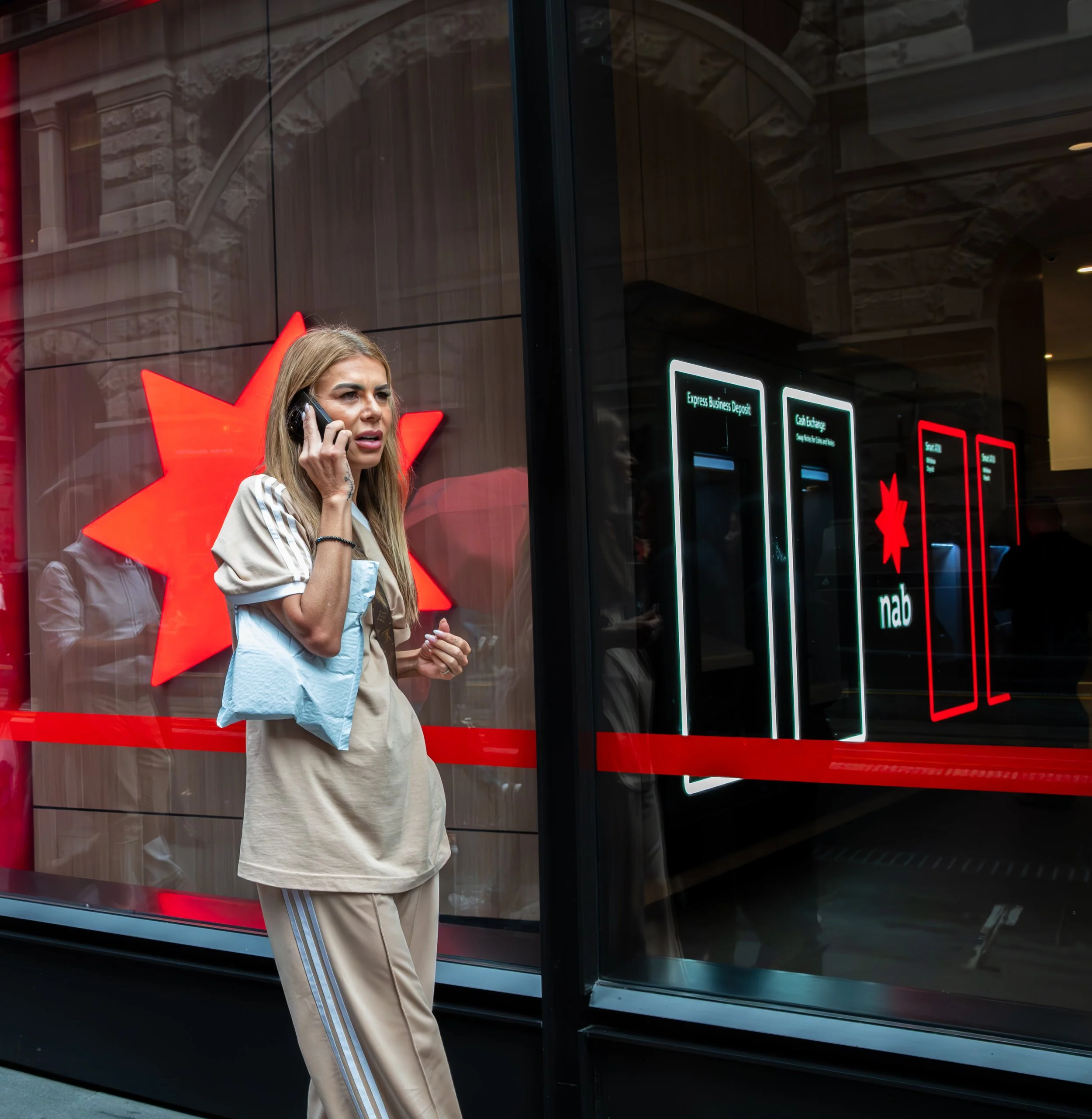A woman with long hair talking on her mobile phone, standing outside a bank branch with digital ATM machines displayed inside the glass window, and the logo of NAB, an Australian bank.