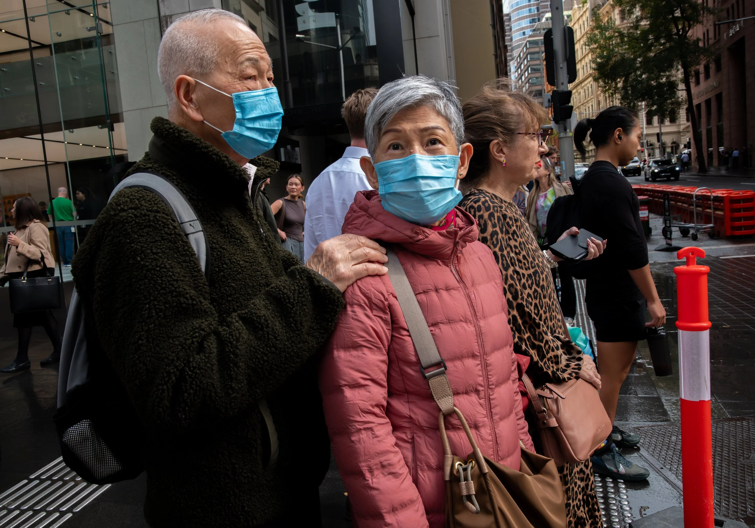 A group of people wearing face masks standing in line on a city sidewalk, with tall buildings and urban scenery in the background.