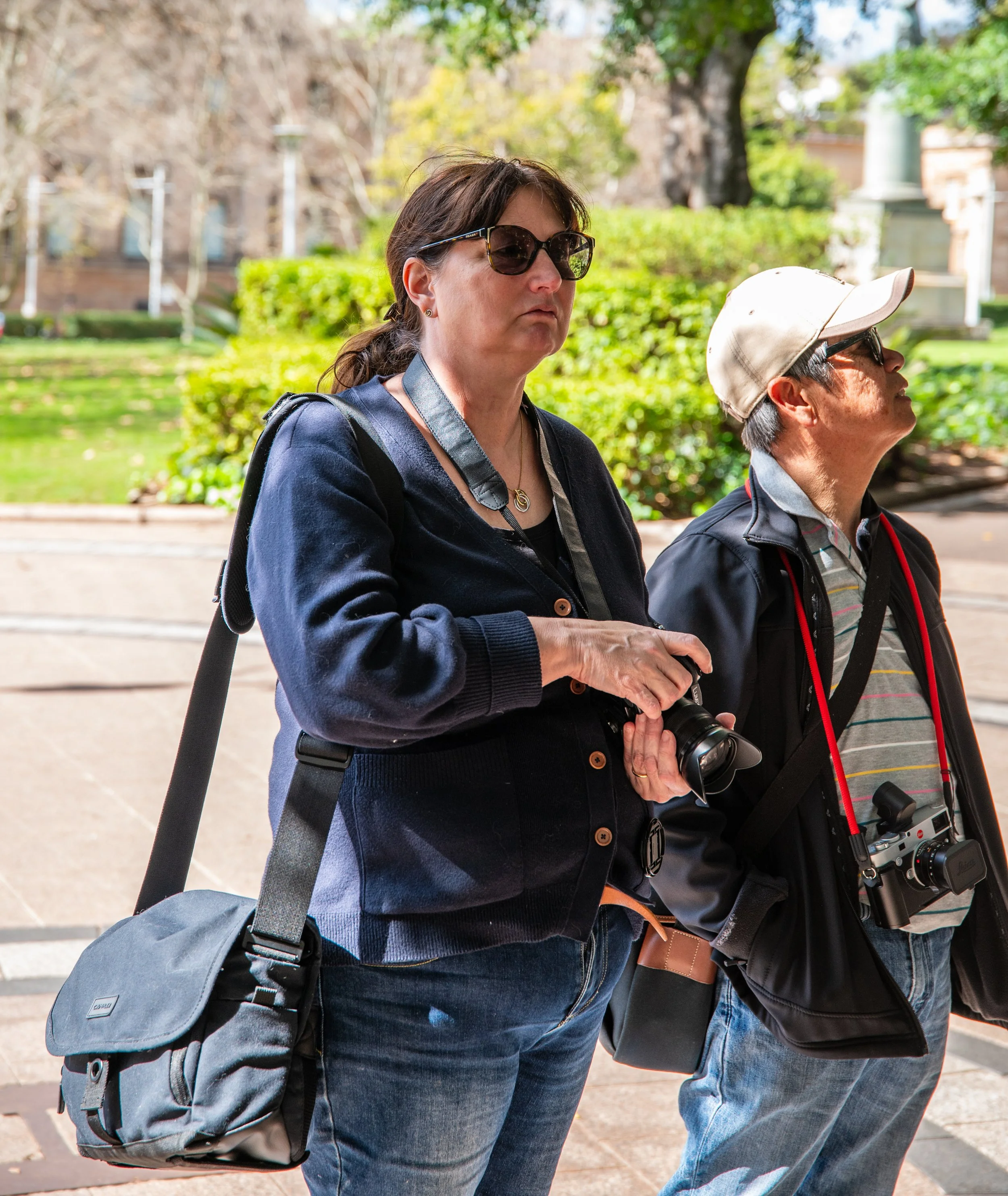 Two middle-aged tourists, a woman and a man, standing outdoors with cameras hanging around their necks. The woman is wearing sunglasses and a navy blue jacket, while the man has a beige cap and a striped shirt. They appear to be sightseeing on a sunny day in a park or historic area.