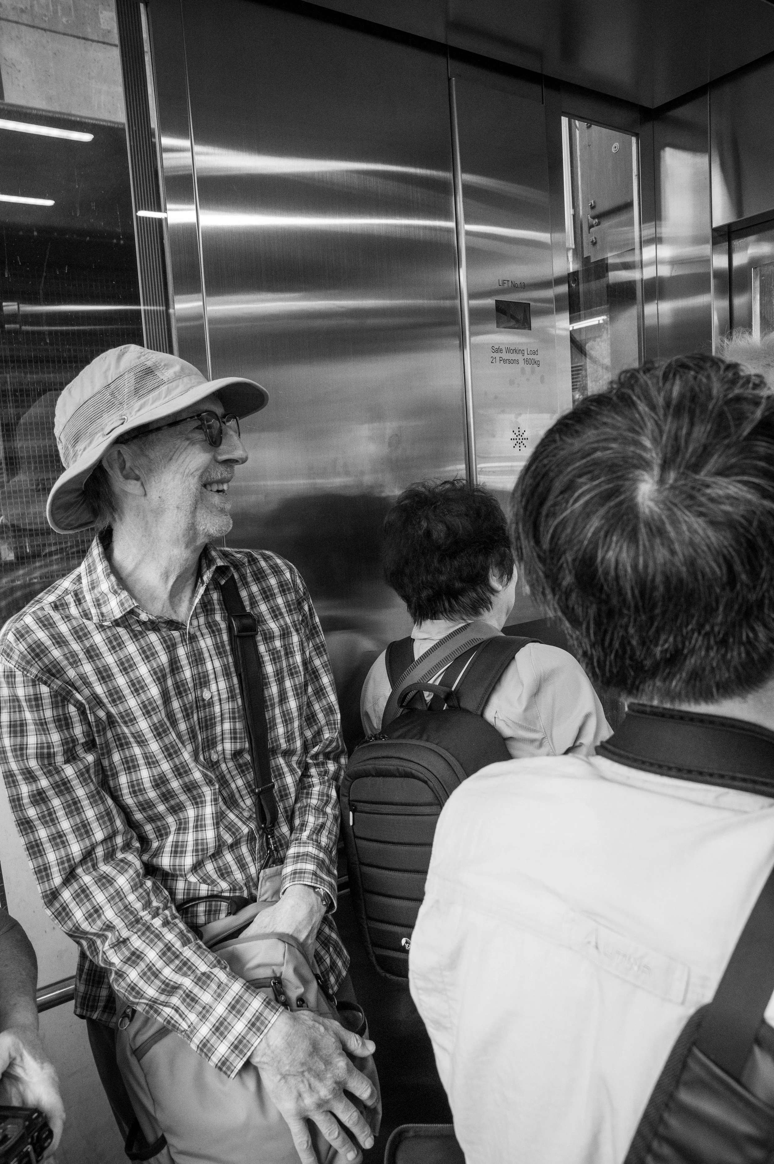 A group of people, including an older man with a hat and glasses, are standing in line inside an elevator, some with backpacks. The elevator has a reflective metal surface and a sign indicating weight capacity.