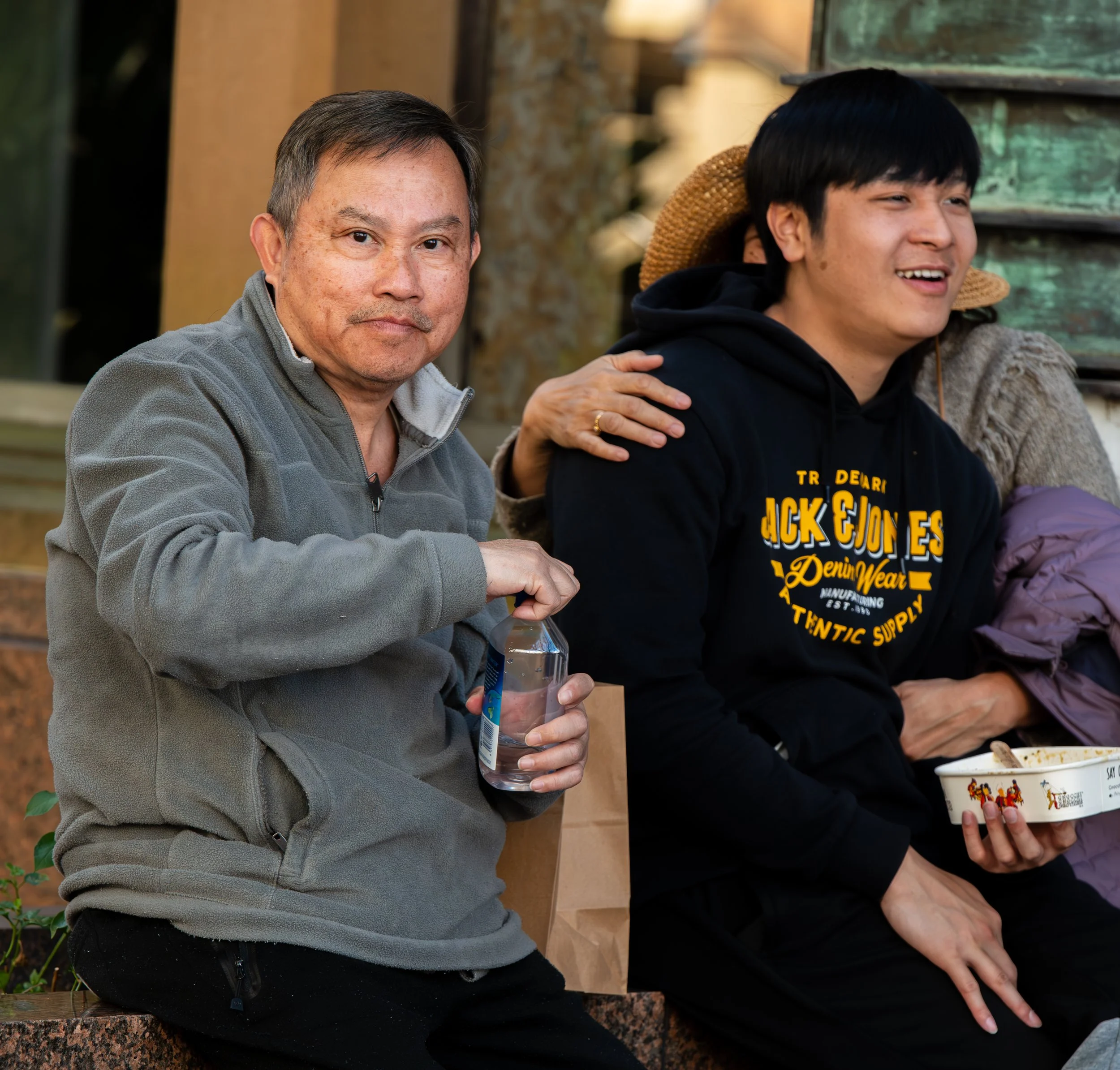 Two men sitting outdoors, one holding a water bottle and the other holding a bowl of food, with friends, smiling and relaxing.