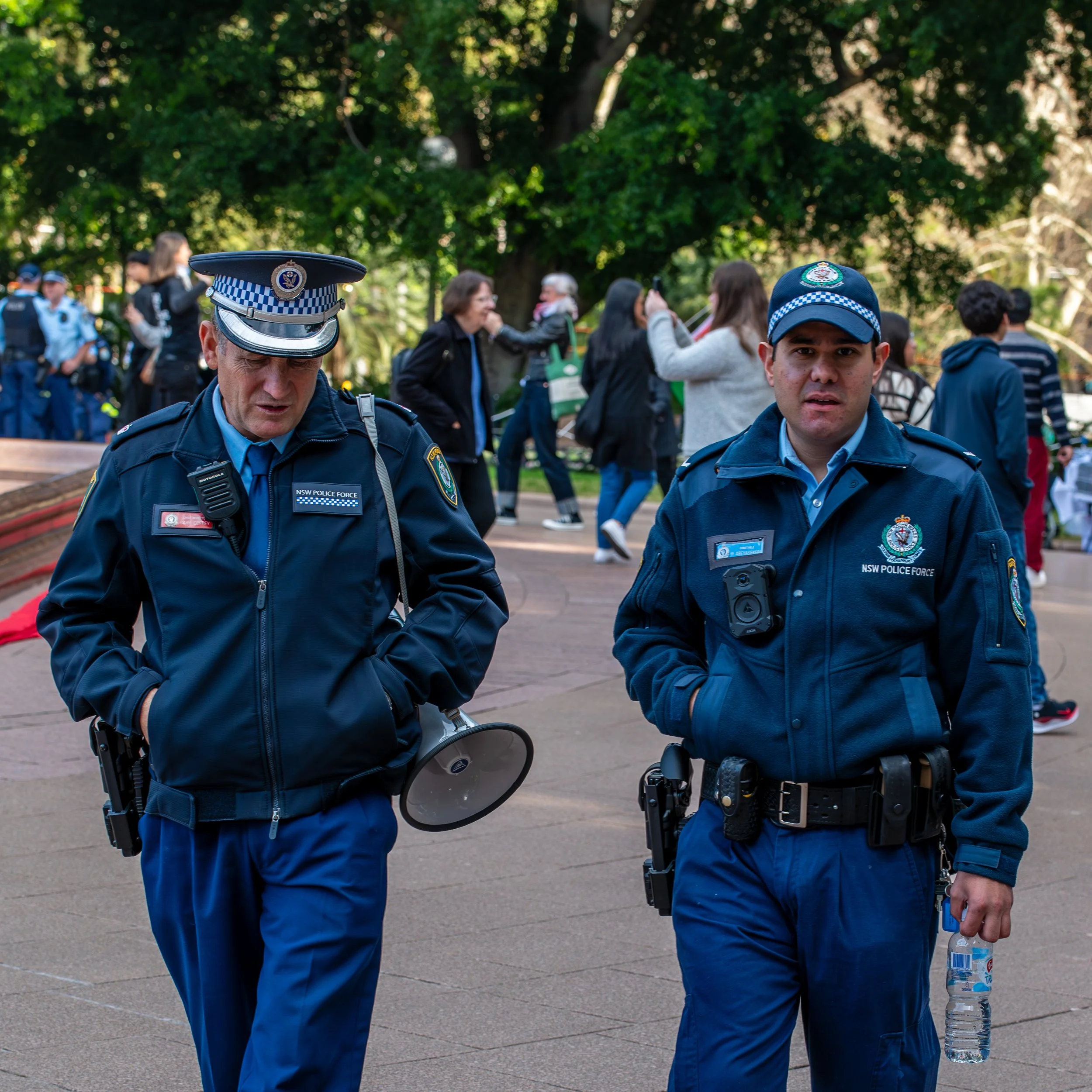 Two police officers in uniform patrolling a park with people walking and taking photos behind them.