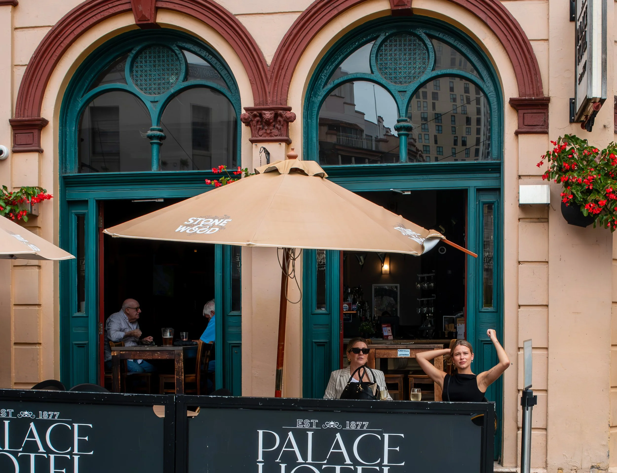 People sitting at an outdoor table of Palace Hotel, with a large beige umbrella overhead, in front of a building with turquoise-trimmed windows and flower boxes.