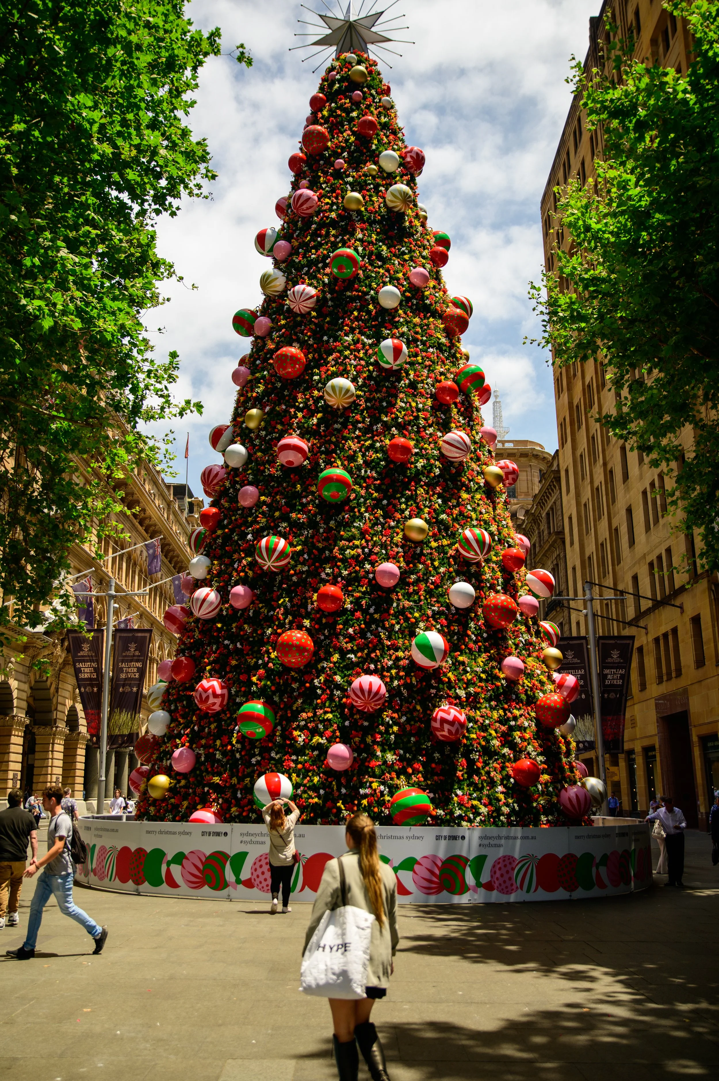 Large decorated Christmas tree with red, green, pink, and gold ornaments, situated in an urban outdoor setting with people walking by and green trees on the sides.