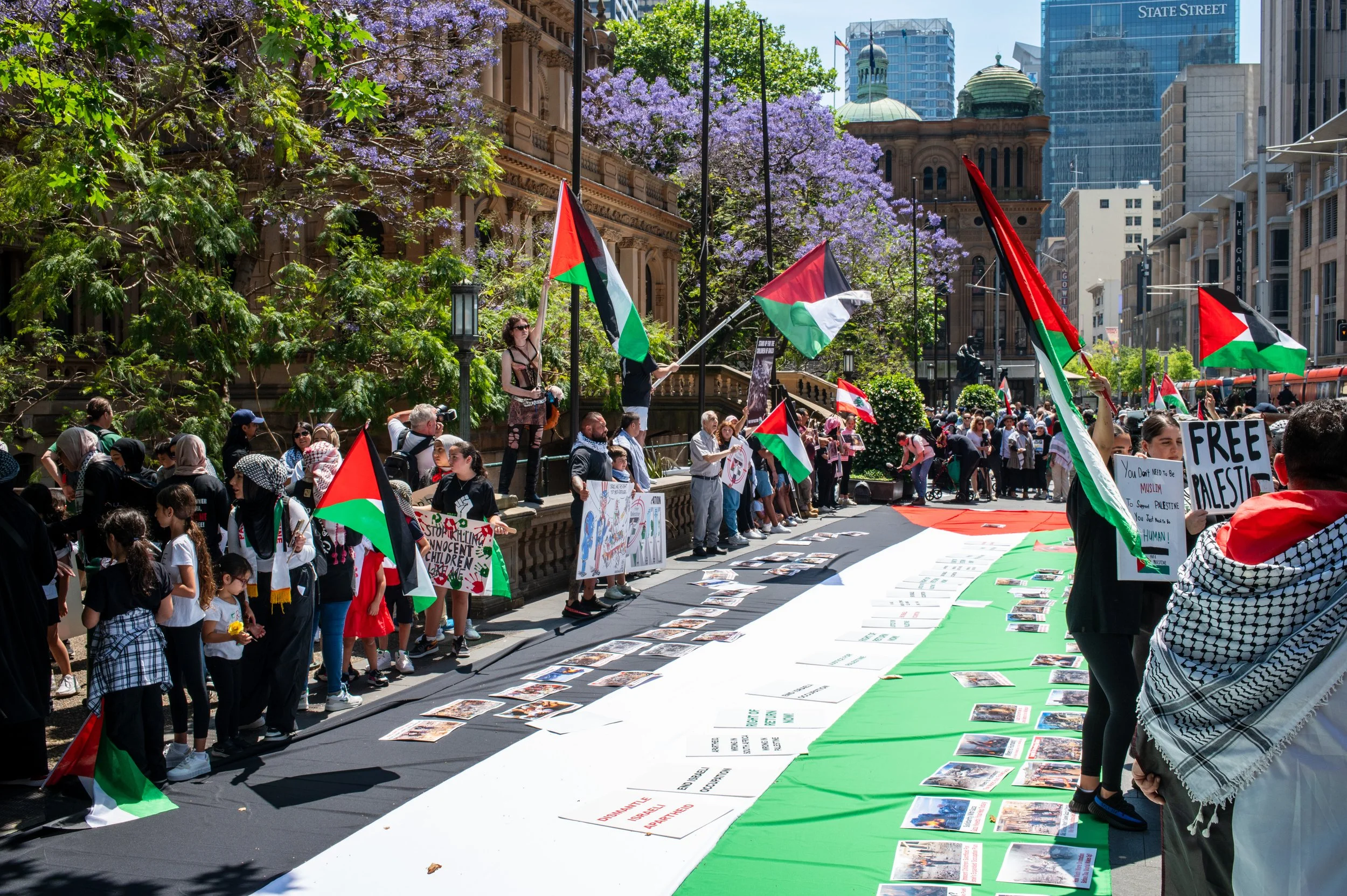 A crowded outdoor protest with people holding Palestinian flags, standing around large signs and banners on a city street. Some signs call for free Palestine and highlight issues in Palestine. The scene includes a large Palestinian flag on the ground as a backdrop, with city buildings and trees with purple flowers in the background.