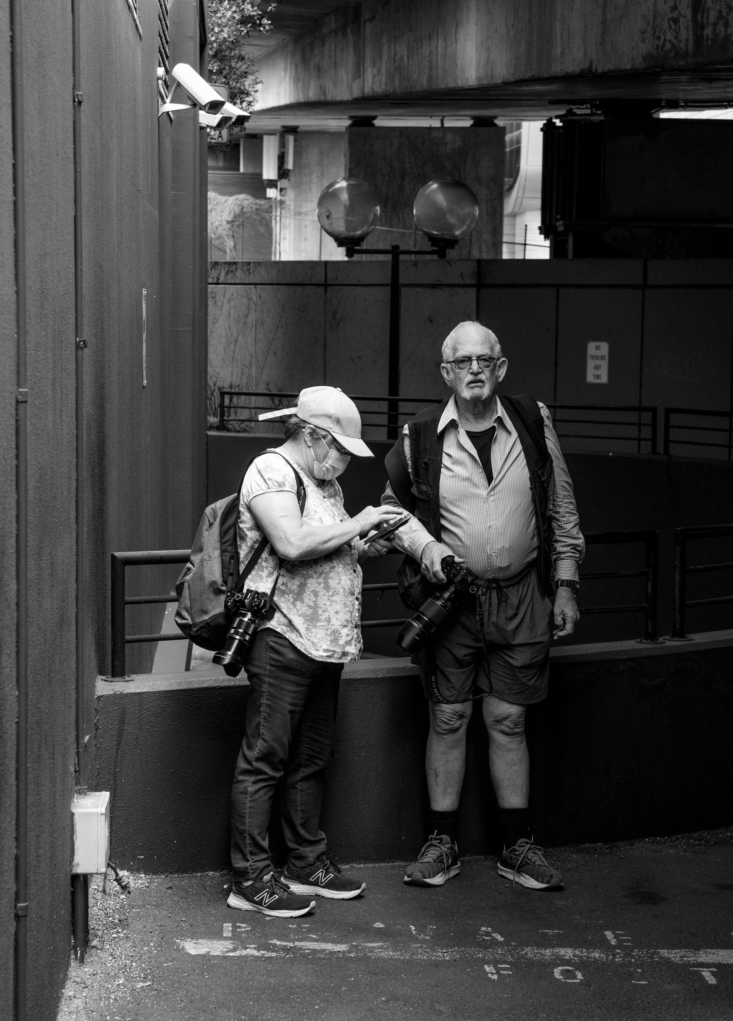 A black-and-white photograph of two people standing on a city sidewalk under an overpass. One person, an older man, has a camera hanging from his neck and is looking directly at the camera. The other person, a woman, is wearing a hat, face mask, and glasses, and is looking at her phone.