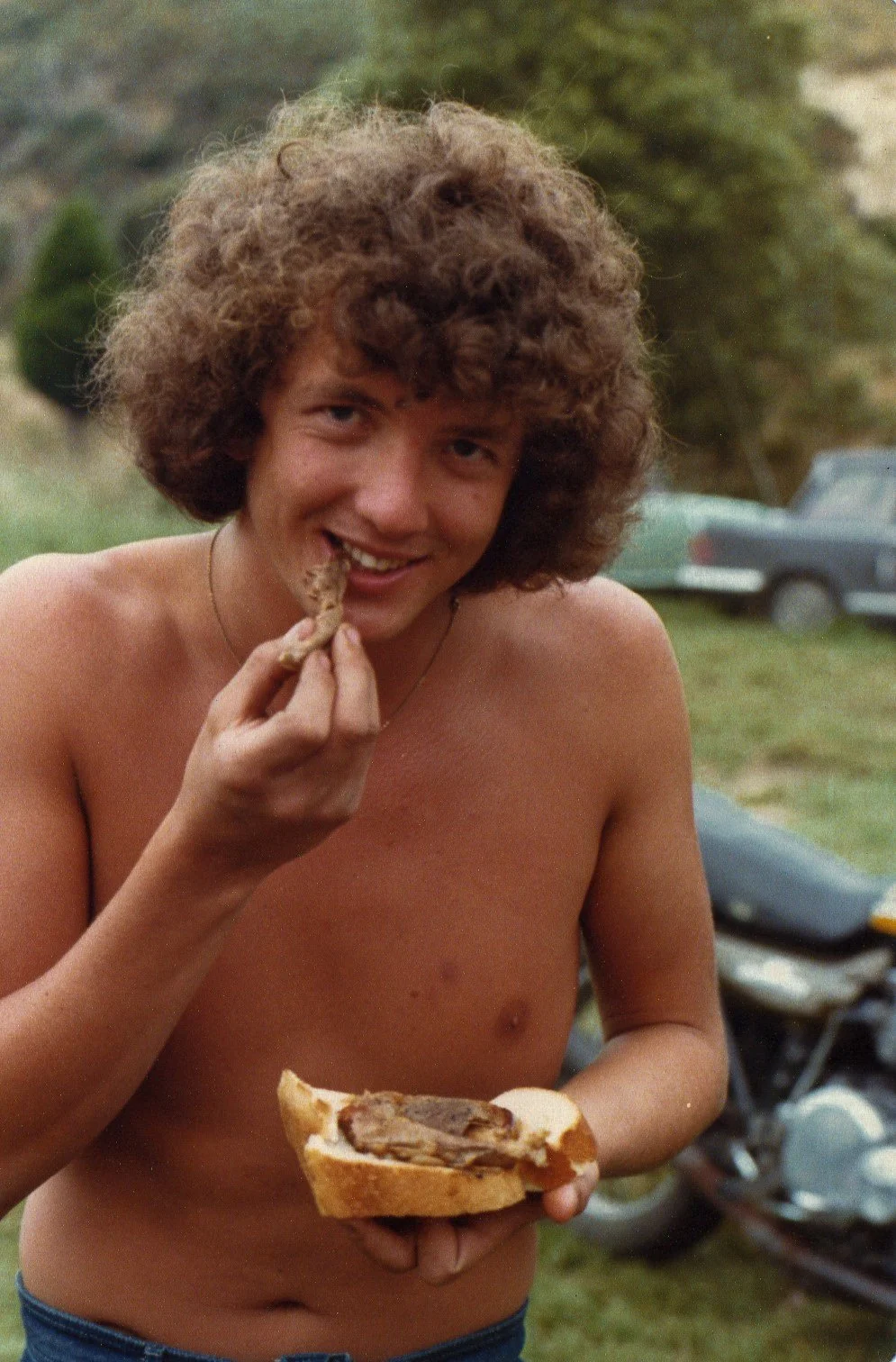 A shirtless young man with curly hair smiling while eating meat from a sandwich outdoors.