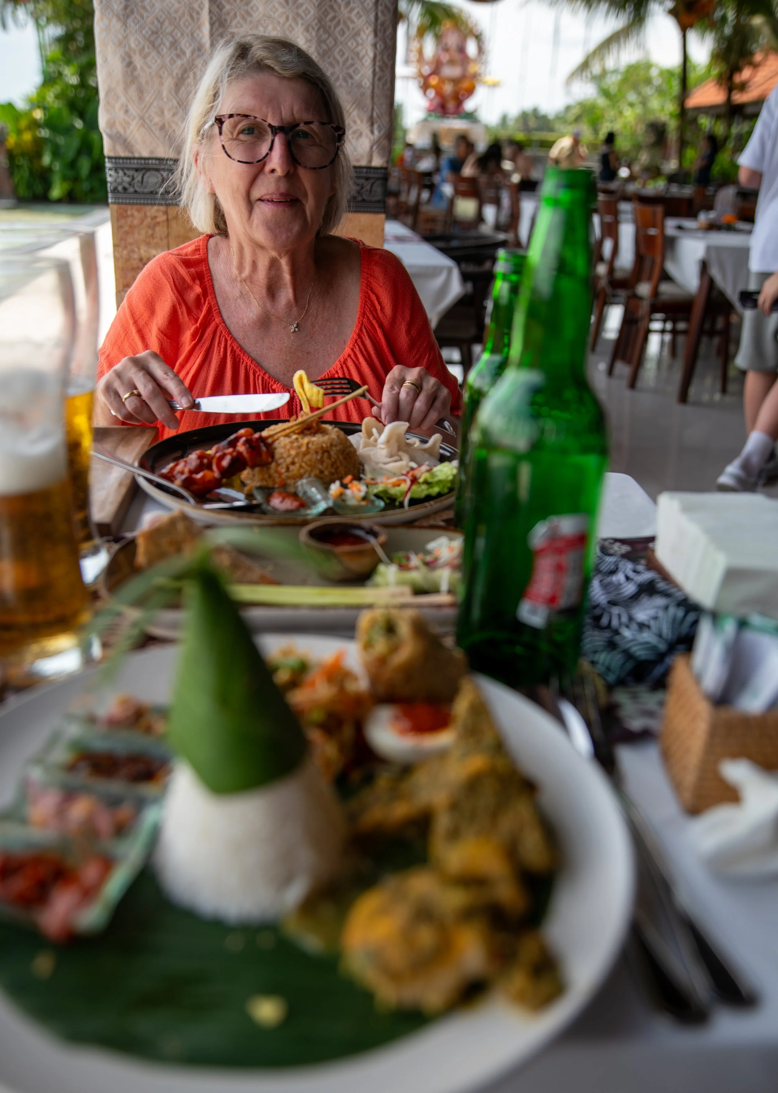 Older woman with glasses wearing an orange top, sitting at a table with a variety of Asian dishes including rice, vegetables, and appetizers in a restaurant with outdoor seating and other diners in the background.