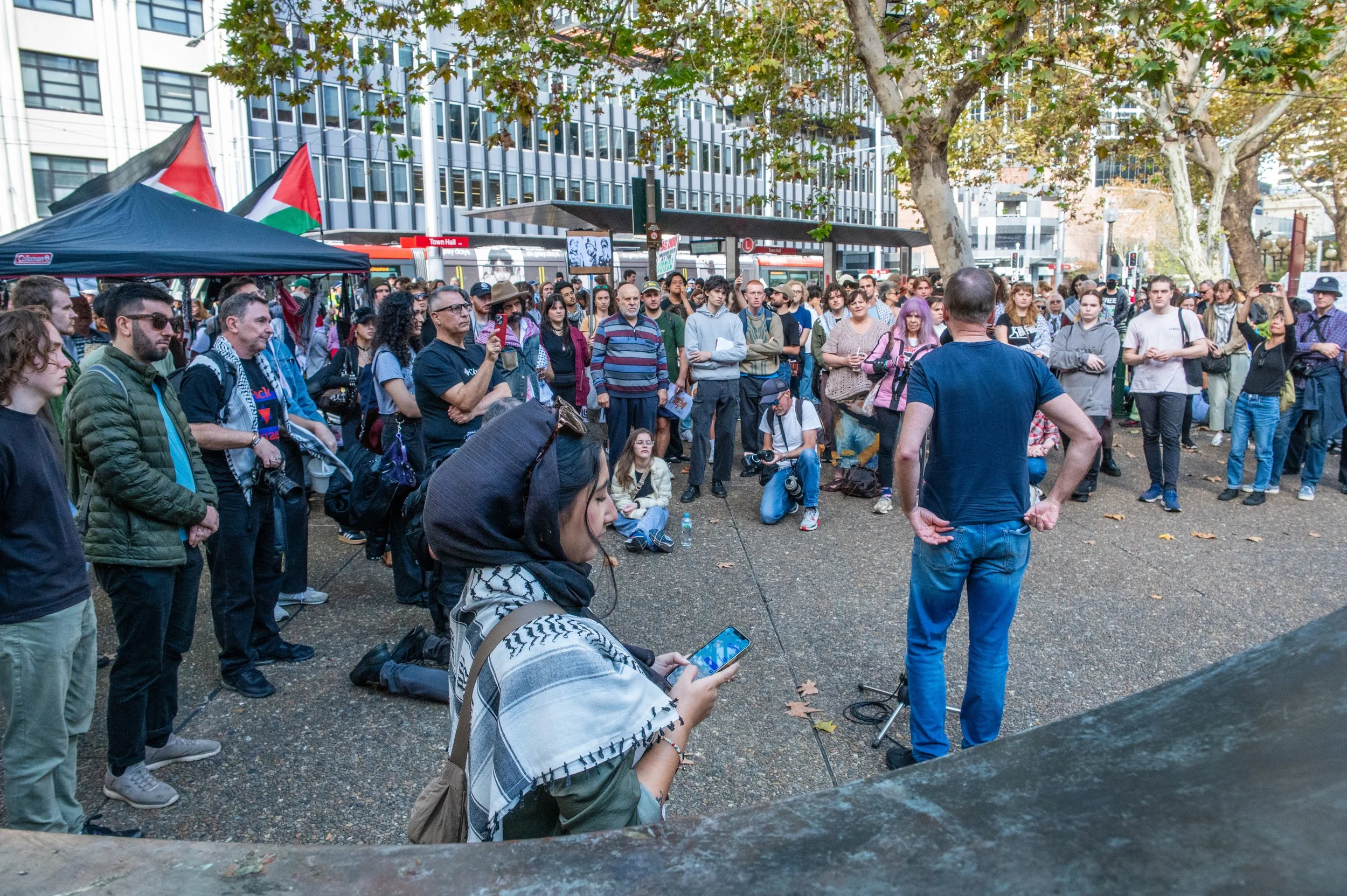 Crowd gathered in a city park, listening to a person speaking at an outdoor event. Some attendees are taking photos or recording, with flags visible in the background.