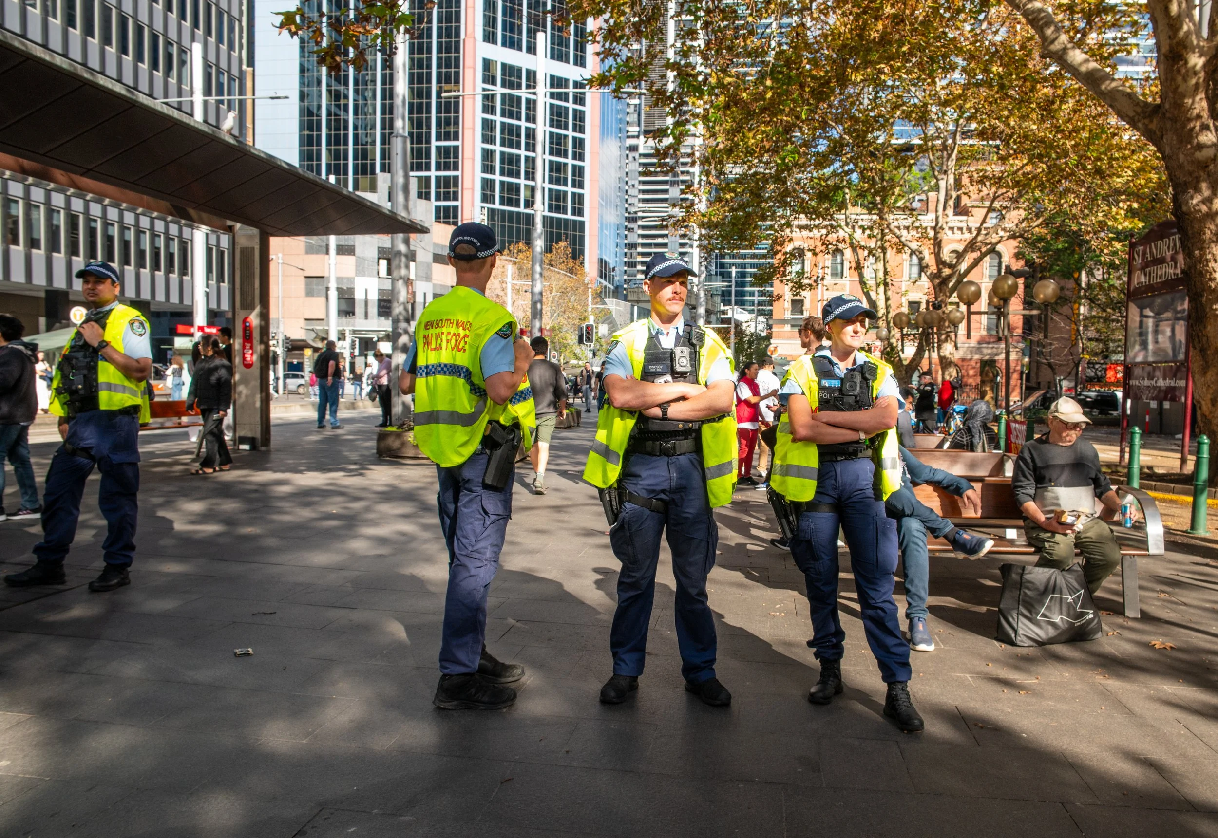 Four police officers in high-visibility vests standing on a city sidewalk, with several people and tall buildings in the background.