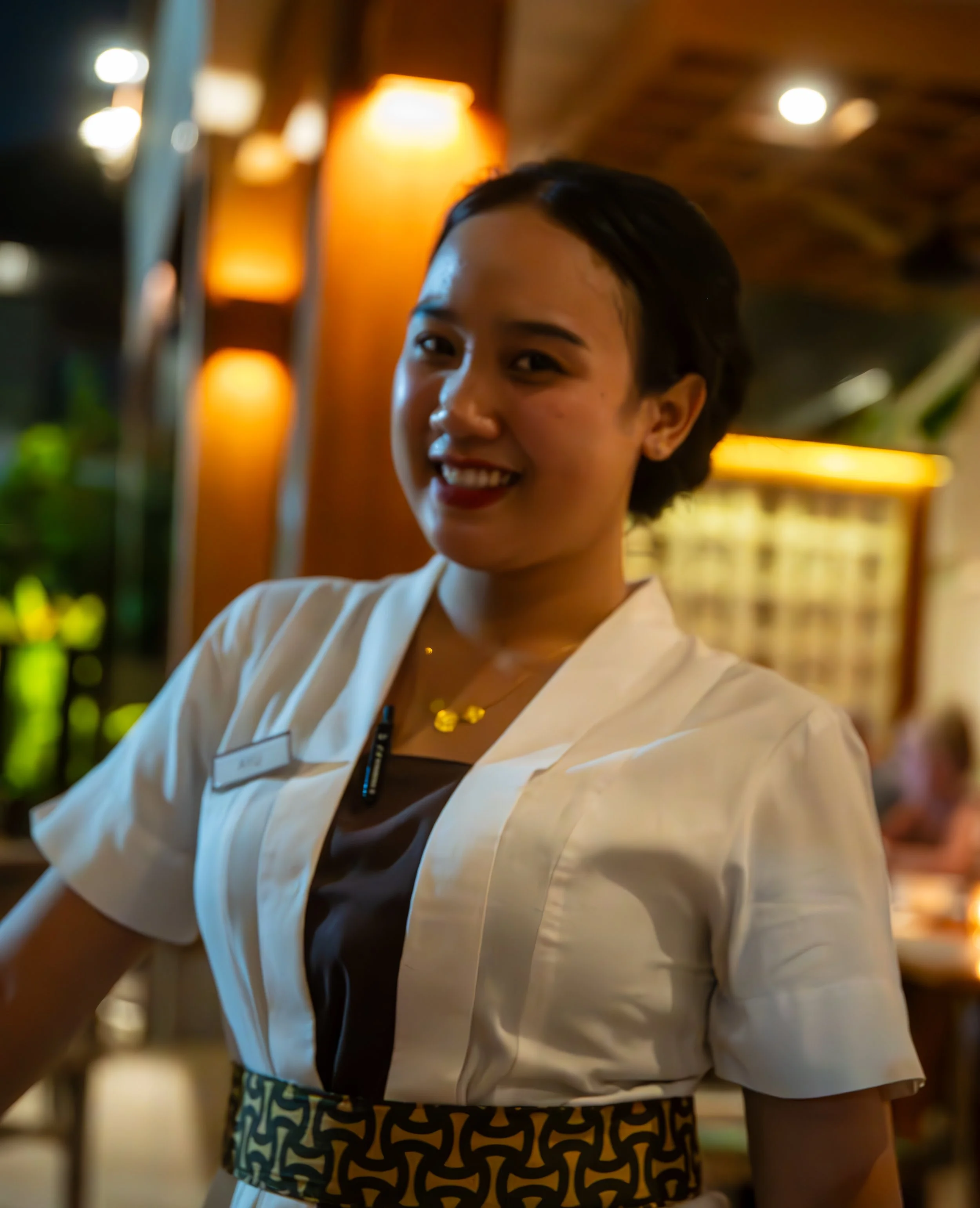 A smiling woman wearing a white uniform with a name tag and a patterned apron, standing in a warmly lit indoor setting with blurred warm-colored lights in the background.