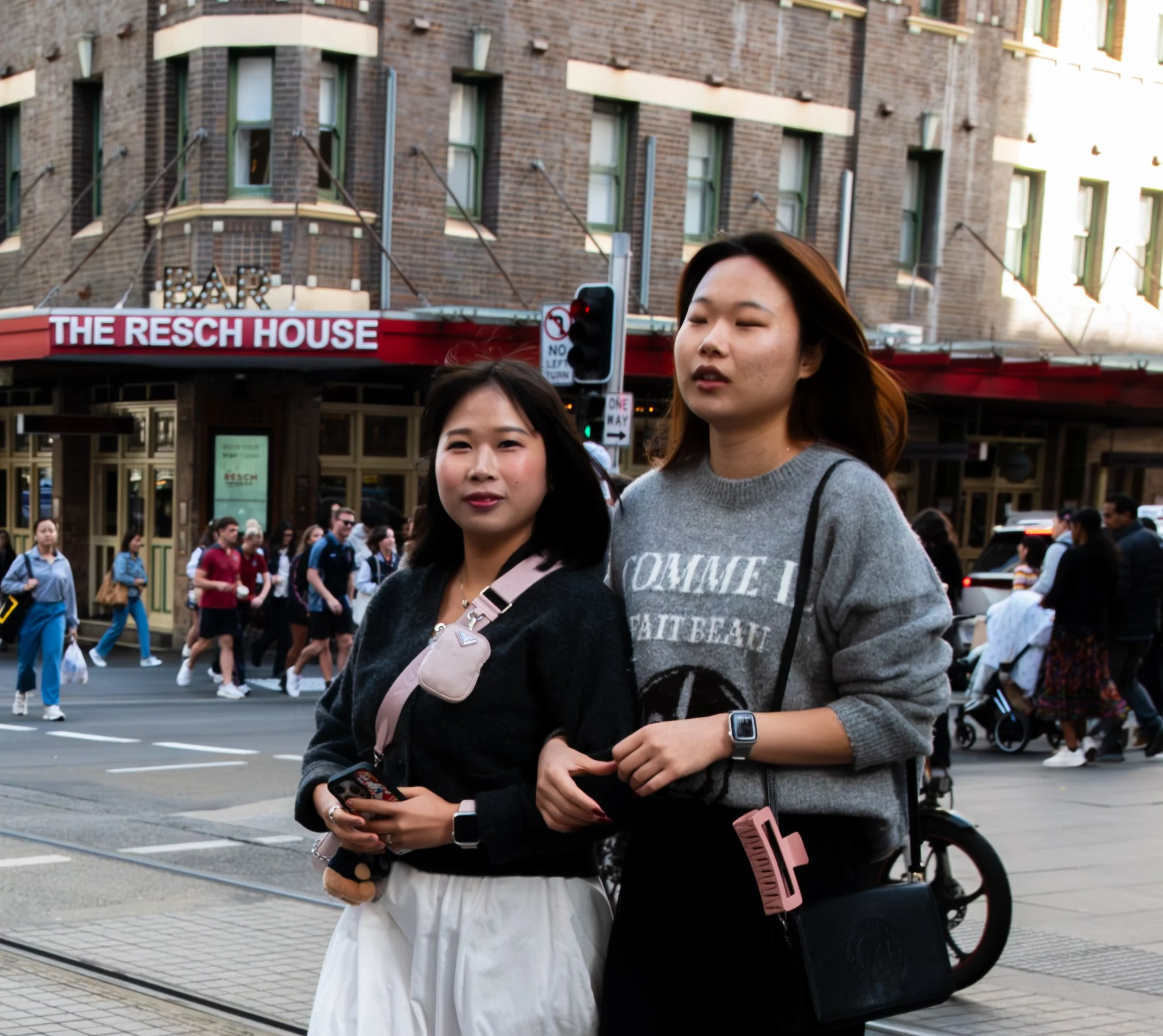 Two young women walk across a busy city street, one with shoulder-length black hair and a pink strap bag, the other with shoulder-length brown hair, wearing a gray sweater, carrying a black bag with a pink item attached, in front of 'The Resch House' restaurant.